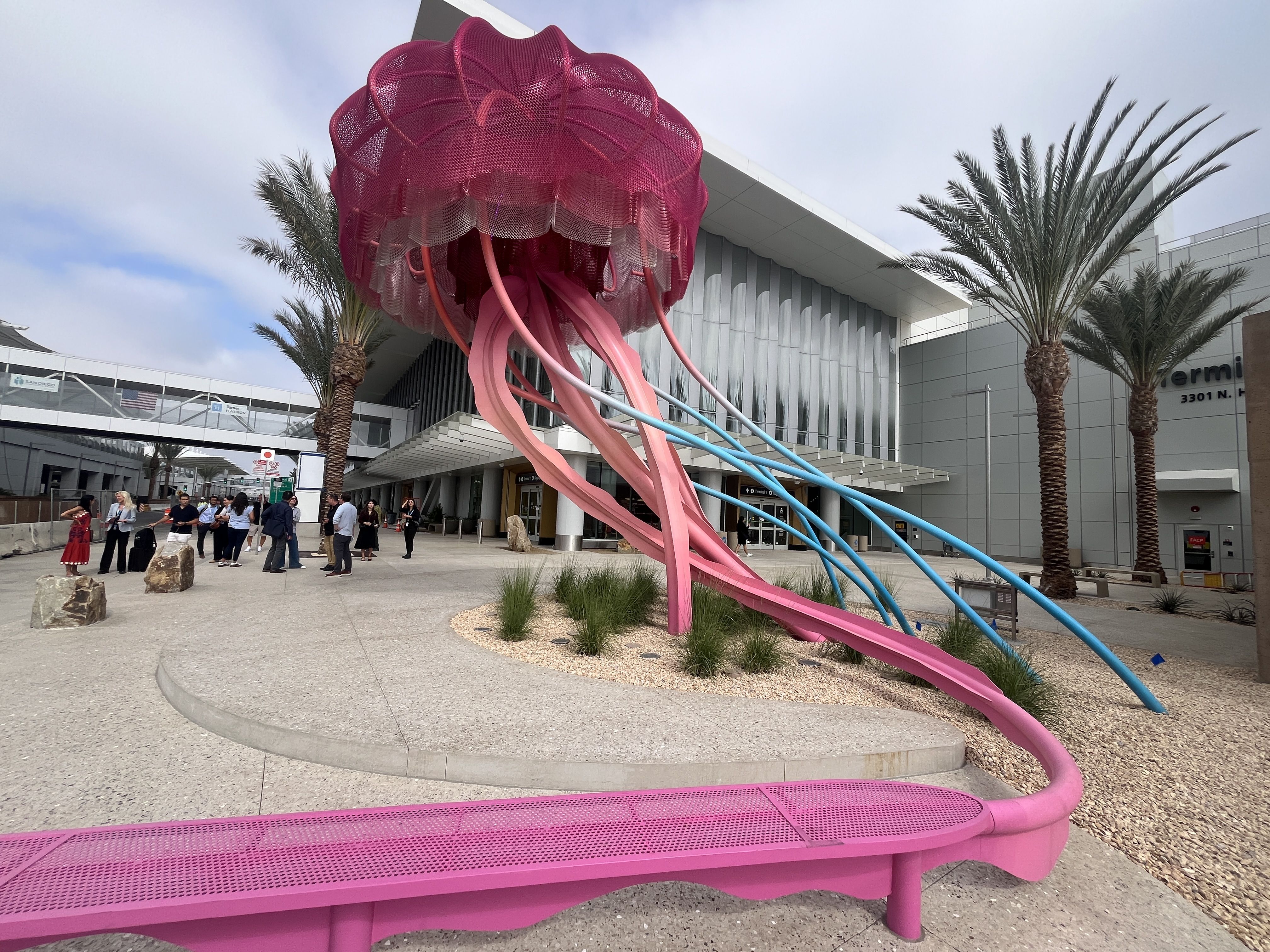 Large pink and blue jellyfish sculpture with long tentacles displayed outside the new Terminal 1 buildings at the San Diego airport with palm trees and people standing nearby.