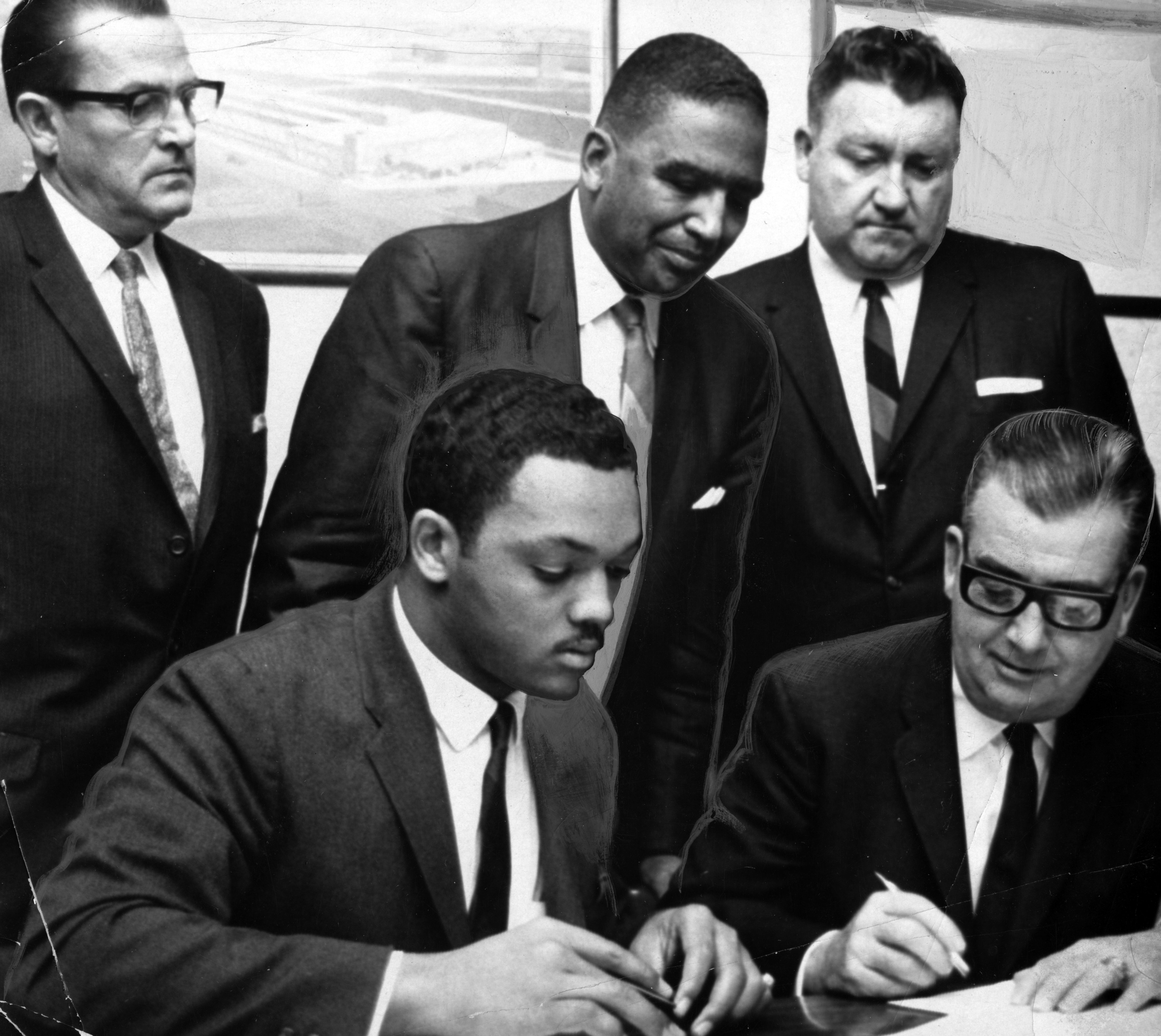 Black and white photo of five men in suits; two seated and signing documents, three standing behind them watching the process intently.