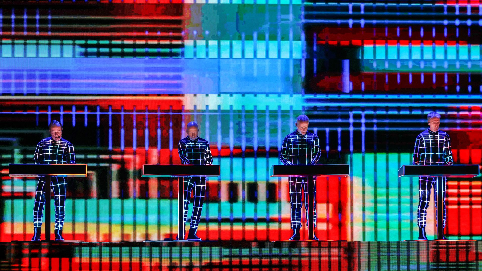 Four individuals wearing suits with illuminated piping play keyboards in front of a projection screen with multi-colored blocks and circles