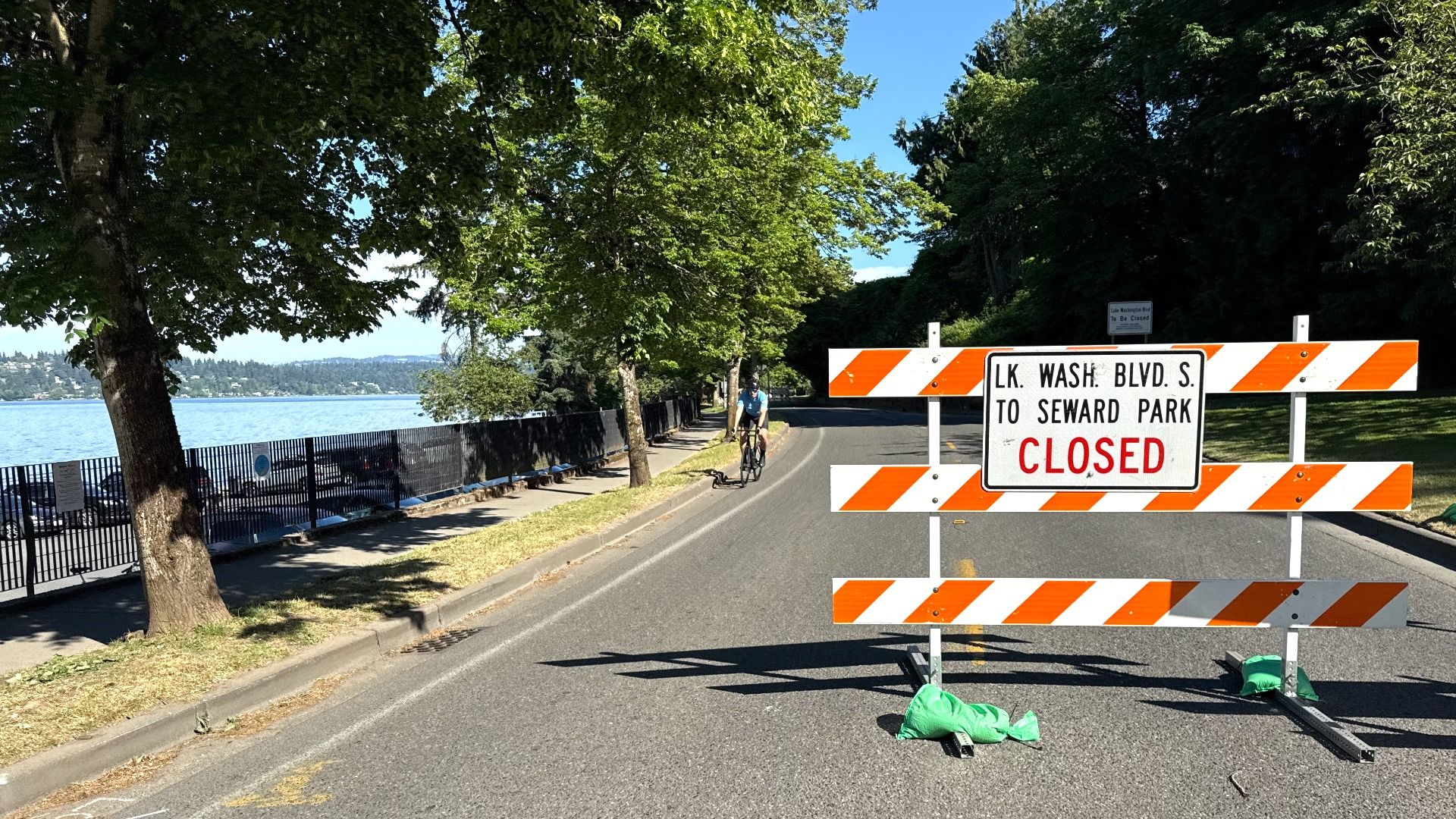 Sunlit lakeside street with trees; an orange-and-white barricade blocks the road. A cyclist rides by a sign reading "LK. WASH. BLVD. S. TO SEWARD PARK" and "CLOSED".