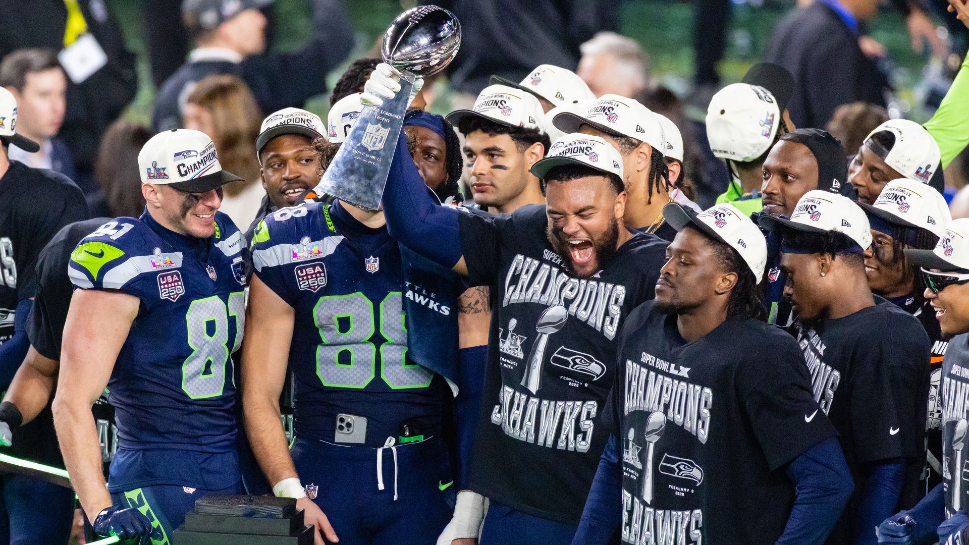 Seattle Seahawks players wearing ball caps and jerseys -- some with shirts that say "Champions Seahawks" -- gather while one player holds up the Vince Lombardi trophy.