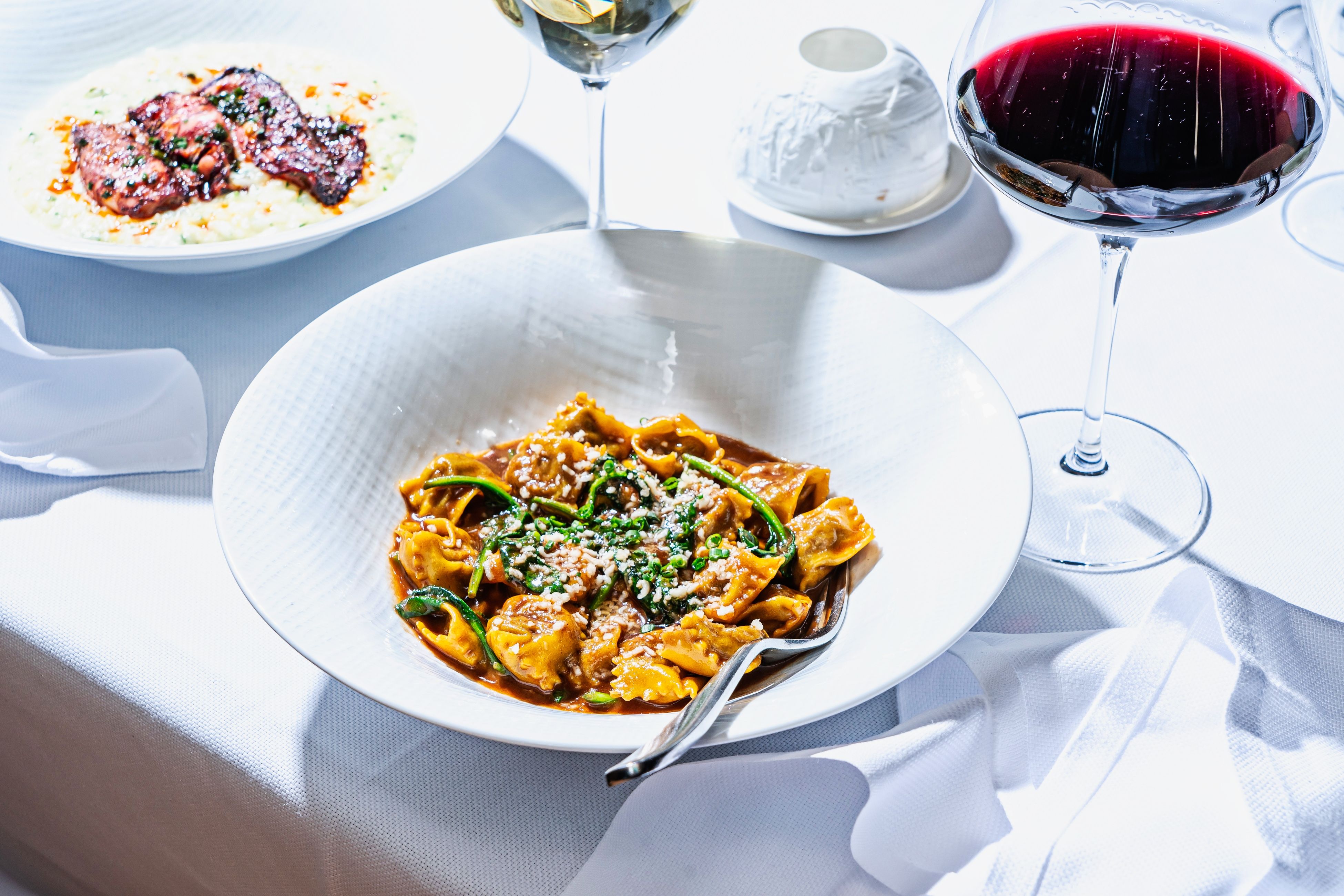 agnolotti with beef short ribs, center, and risotto with wild calamari and a glass of red wine - all on a white tablecloth.