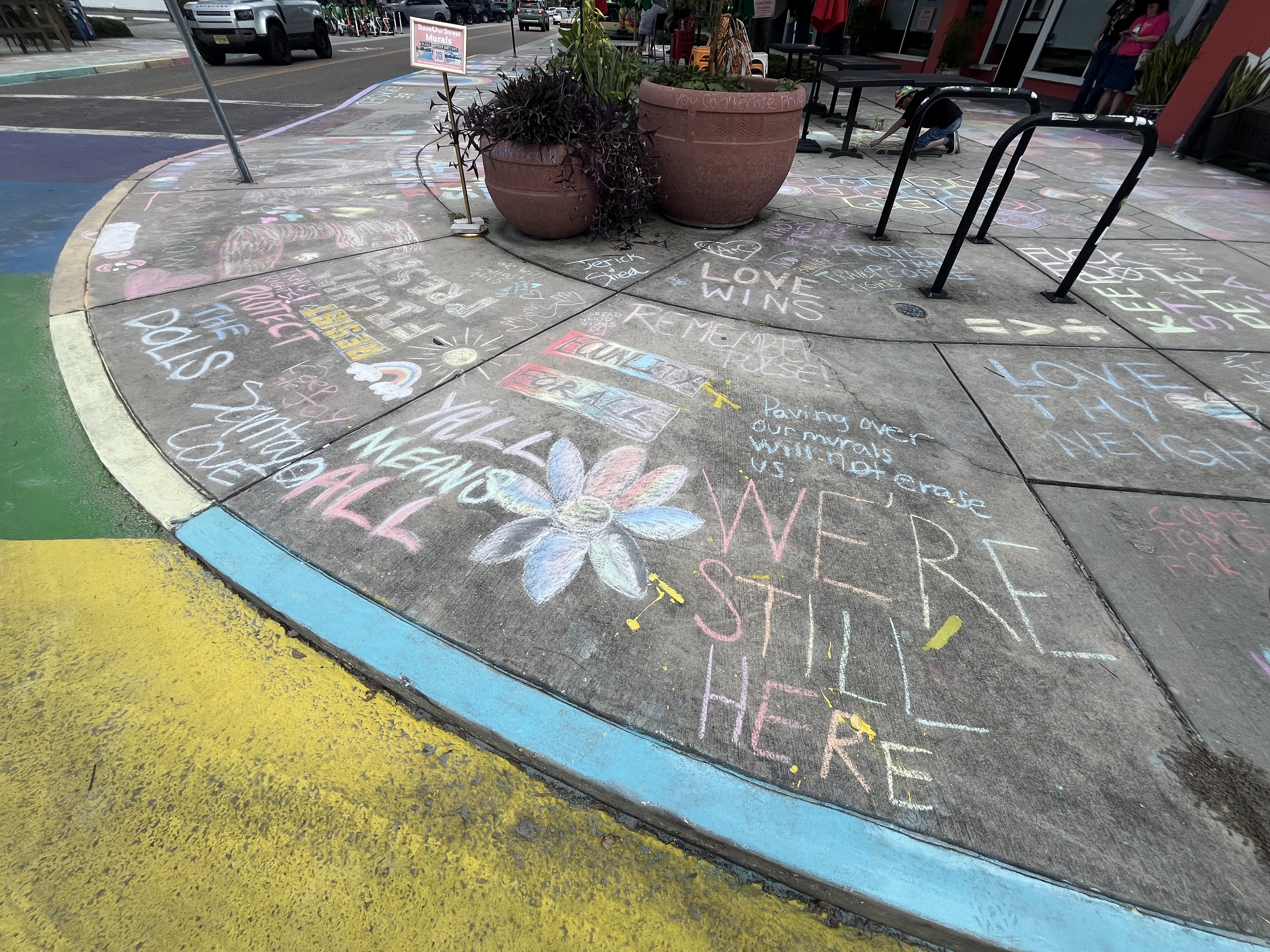 Sidewalk chalk art on pavement includes messages like "WE'RE STILL HERE," "LOVE WINS," a large flower, rainbow, and phrases about protecting rights and unity, with colorful borders nearby.