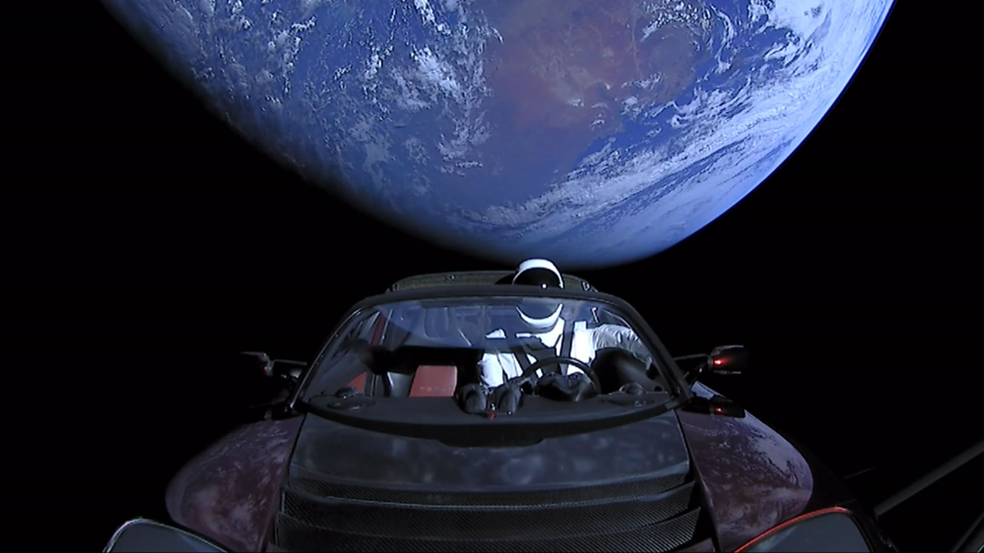 A white-suited astronaut mannequin sits in a red convertible car floating in space with Earth visible in the background against a black sky.