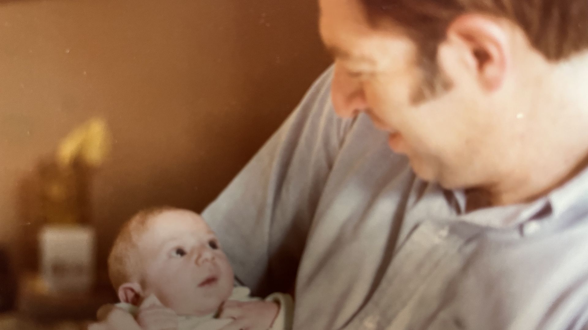 Man in a light blue shirt gently holds a newborn baby, looking down with a soft smile. Warm, dim indoor lighting with a blurred background.