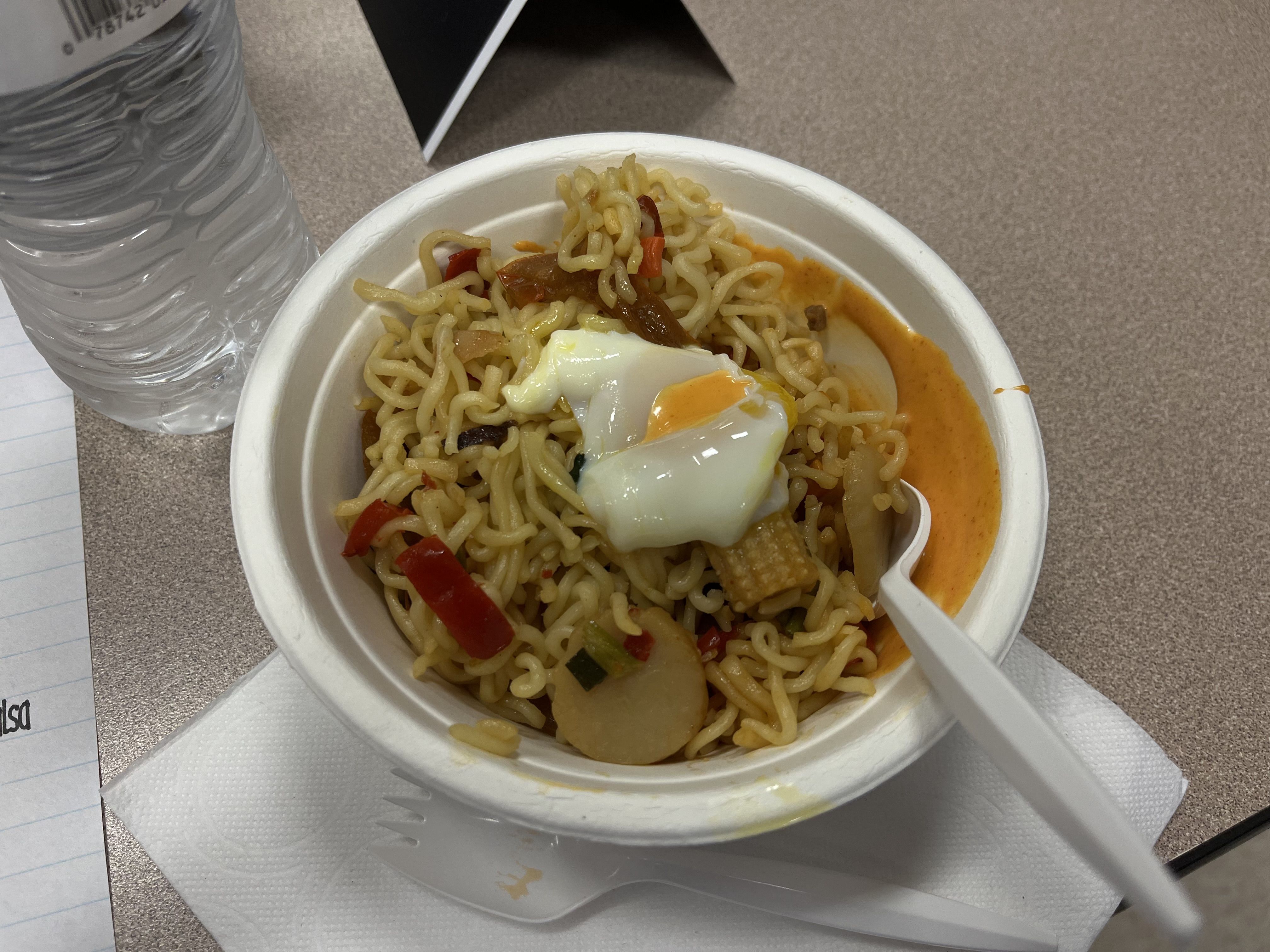 Bowl of noodles with vegetables and a poached egg on top, a plastic spork inside, placed on a white napkin on a table next to a bottle of water and a notepad.