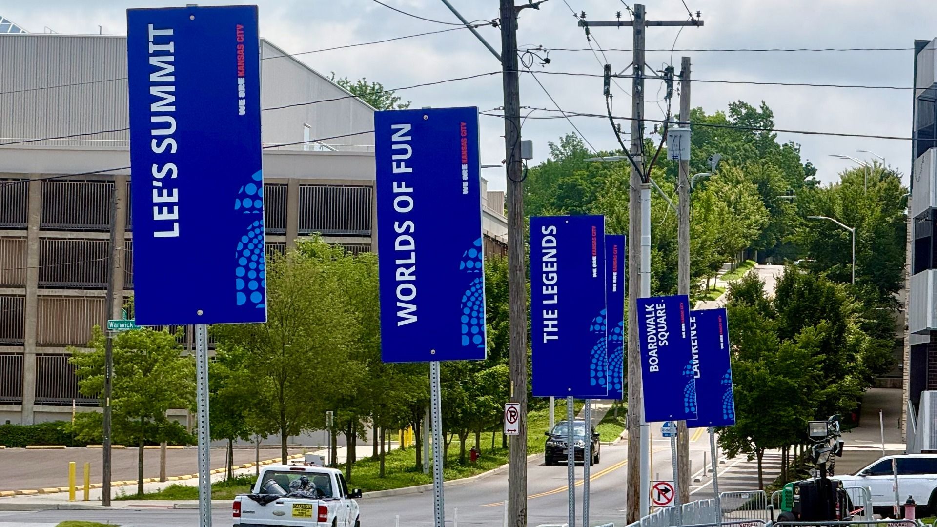 Row of tall blue banners along a street displaying Lees Summit, Worlds of Fun, The Legends, and Boardwalk Square, with power lines, green trees, cars, and a building on the left.