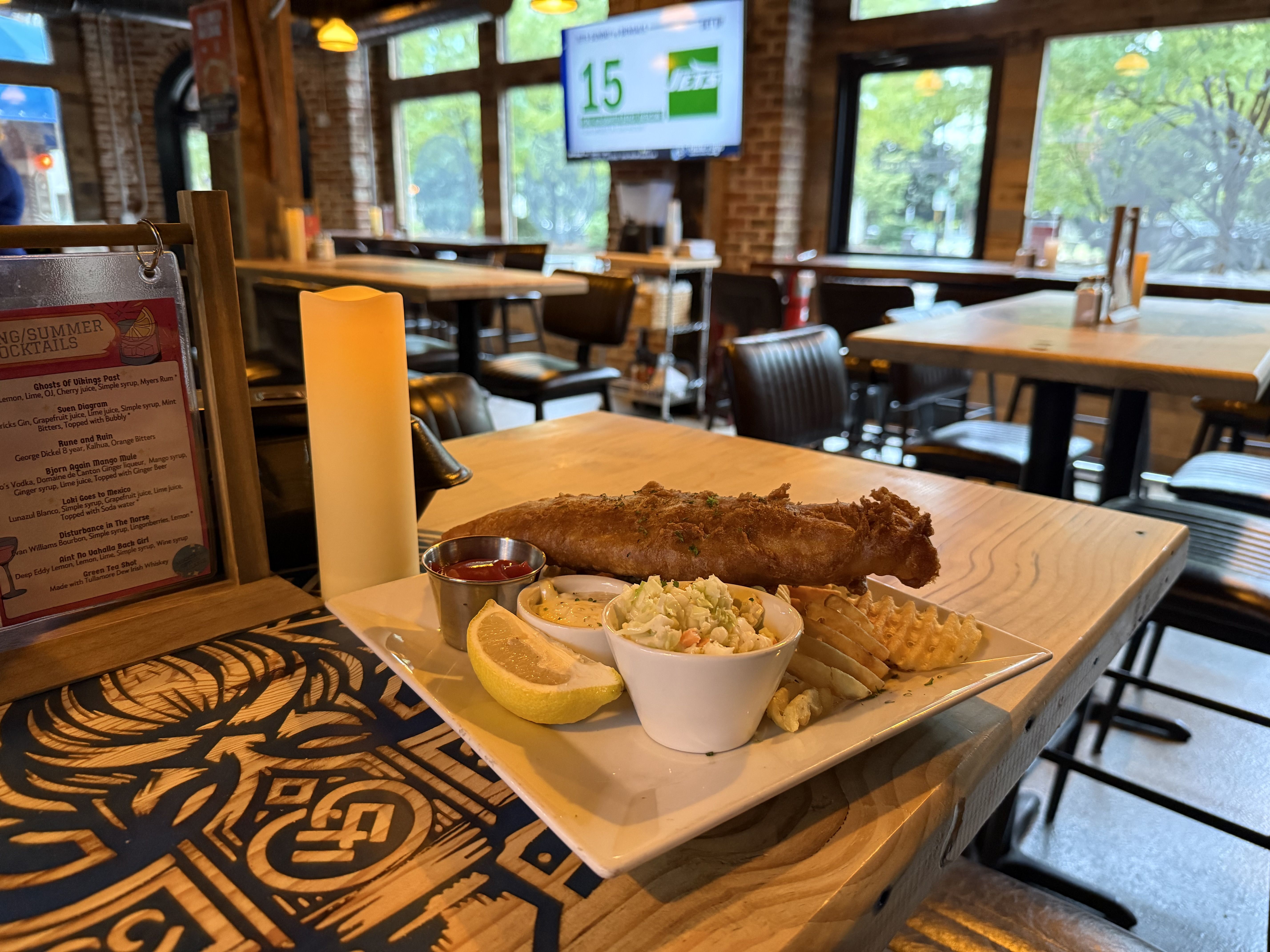 Plate of fried fish, waffle fries, coleslaw, lemon wedge, and sauce on a wooden table in a cozy restaurant with brick walls and large windows showing green trees outside.