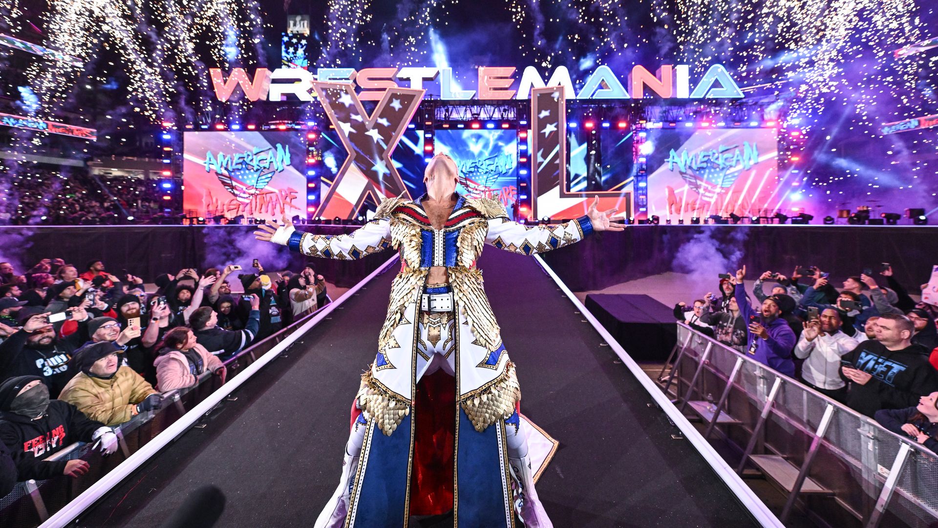 A performer wearing a red, white, blue and gold outfit leans back as a crowd cheers and confetti falls. Behind them is a large Wrestlemania XL logo.