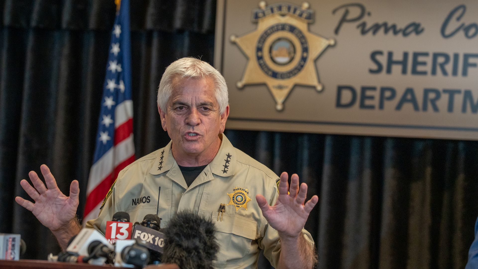 A sheriff in tan uniform with "NANOS" patch speaks at a podium with many microphones in front, an American flag behind him, and a blurred Pima County Sheriff's Department sign in the background.