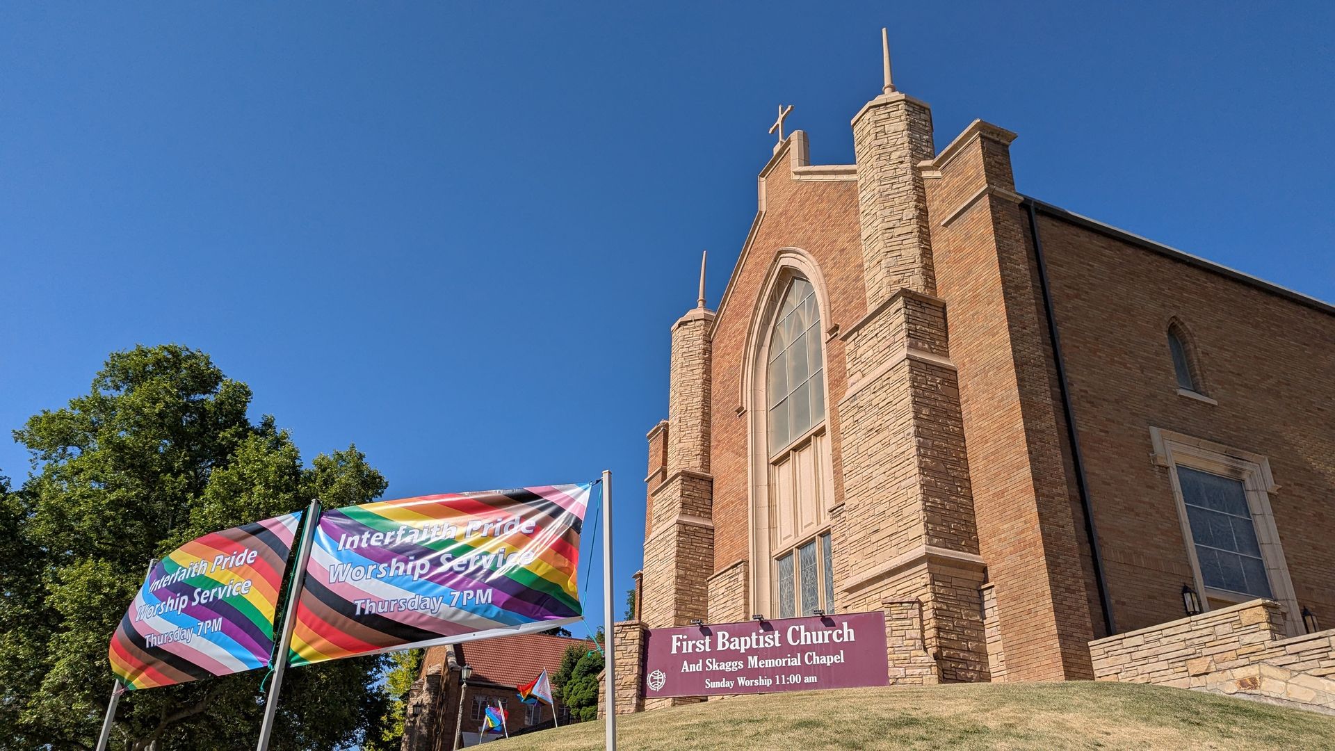 A brick church decorated with pride flags and a sign that reads "First Baptist Church" and a rainbow banner that reads "Interfaith Worship Service Thursday 7pm" 