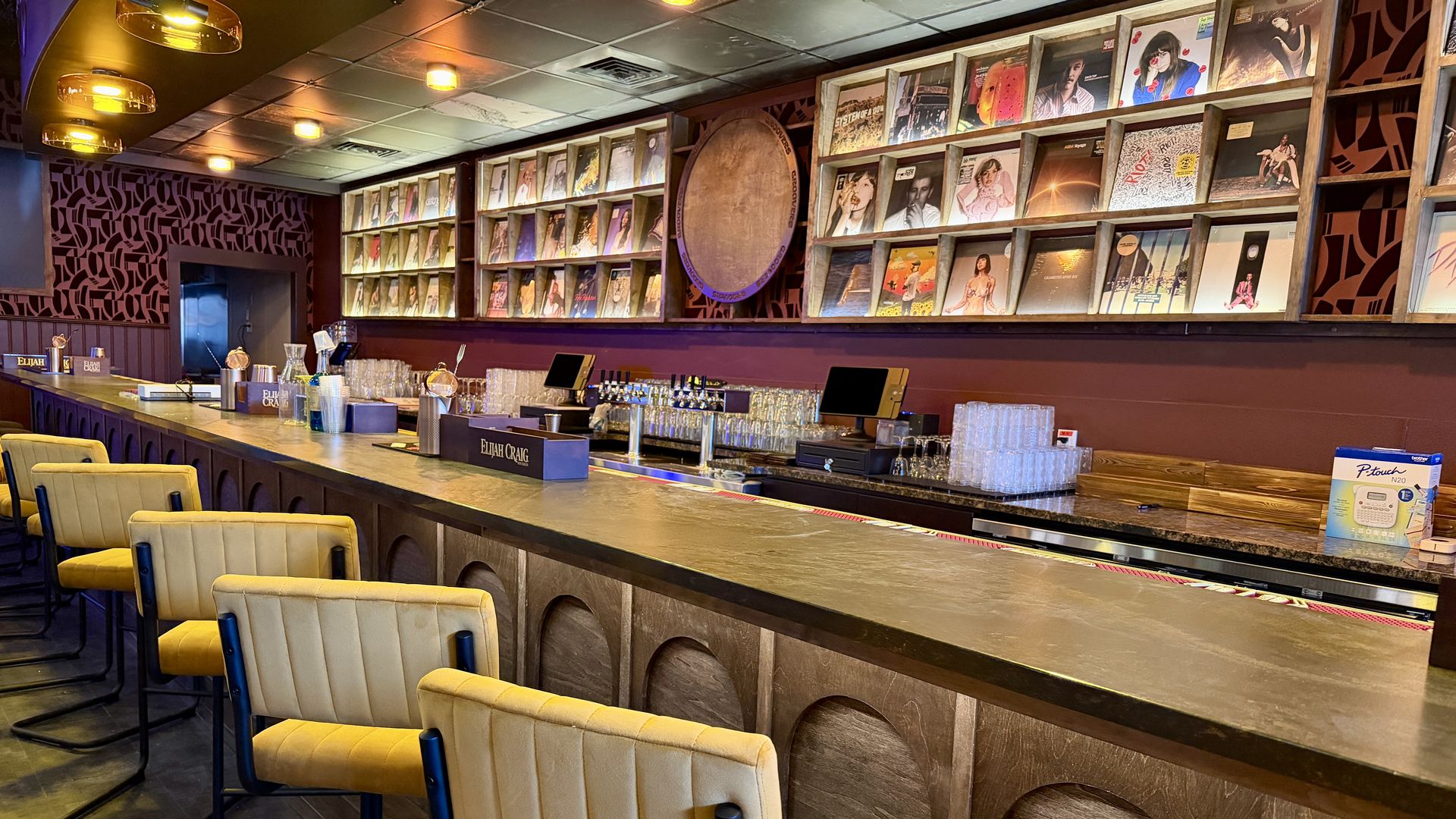 Empty modern bar with mustard yellow padded chairs, wooden counter, shelves displaying vinyl records, warm ambient lighting, and a dark red and brown color scheme.