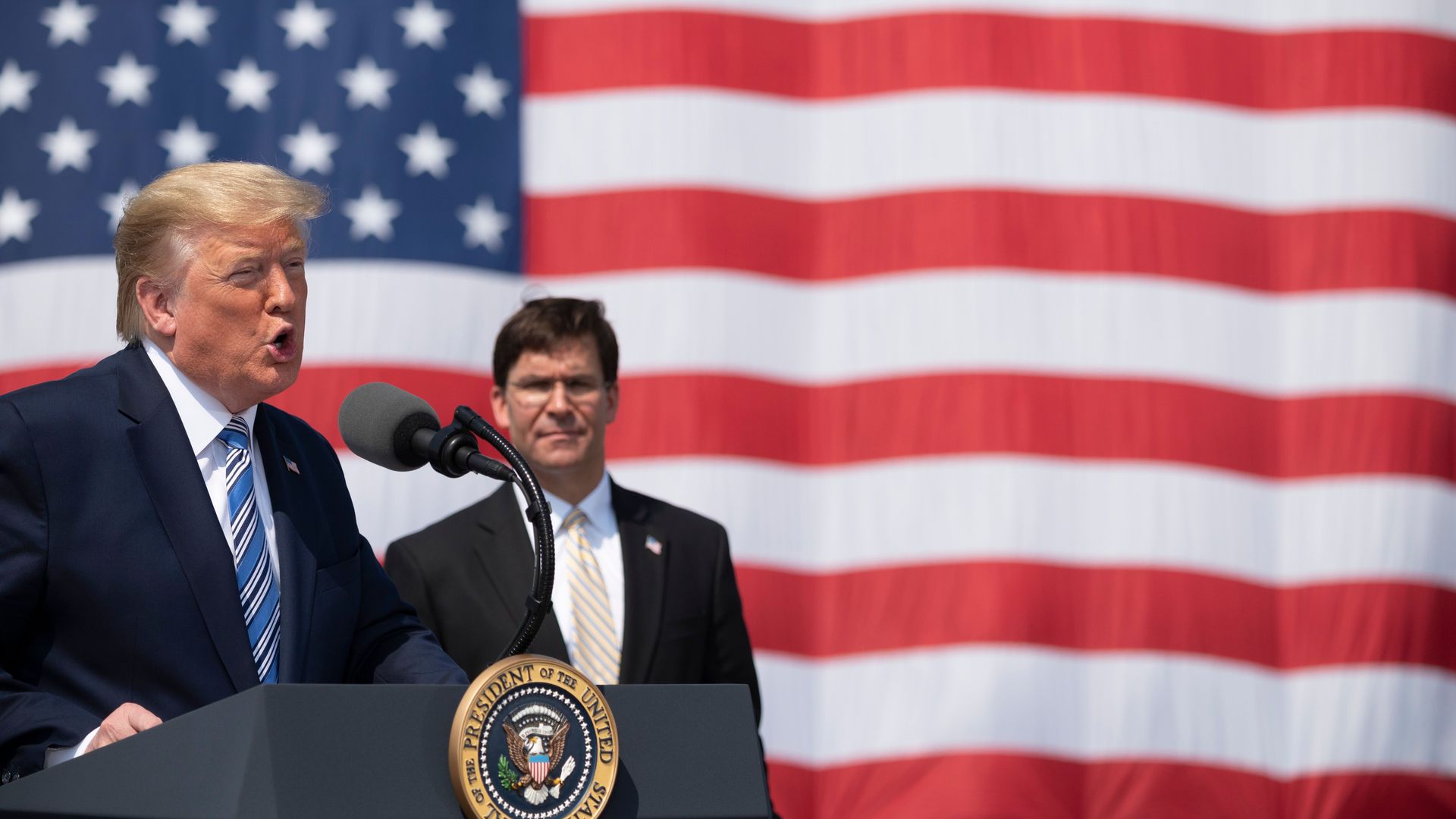 Photo of Donald Trump speaking from a podium as Mark Esper stands behind him against the backdrop of a large American flag