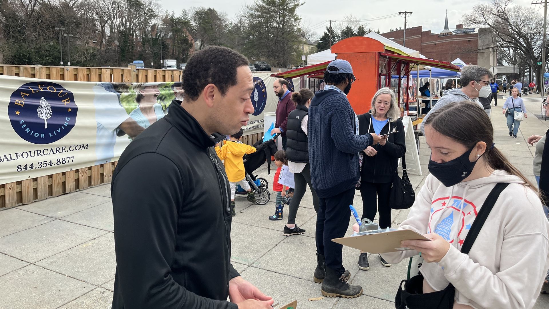 A voter signs his signature for a candidate running for office on the sidewalk at a farmer's market.