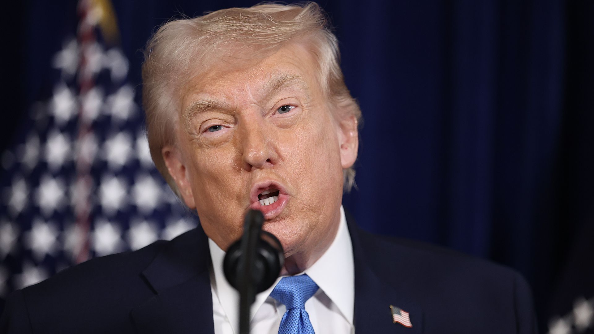 Close-up of President Trump, with gray hair and an American flag pin on dark suit, speaking into a microphone with an American flag blurred in the background.