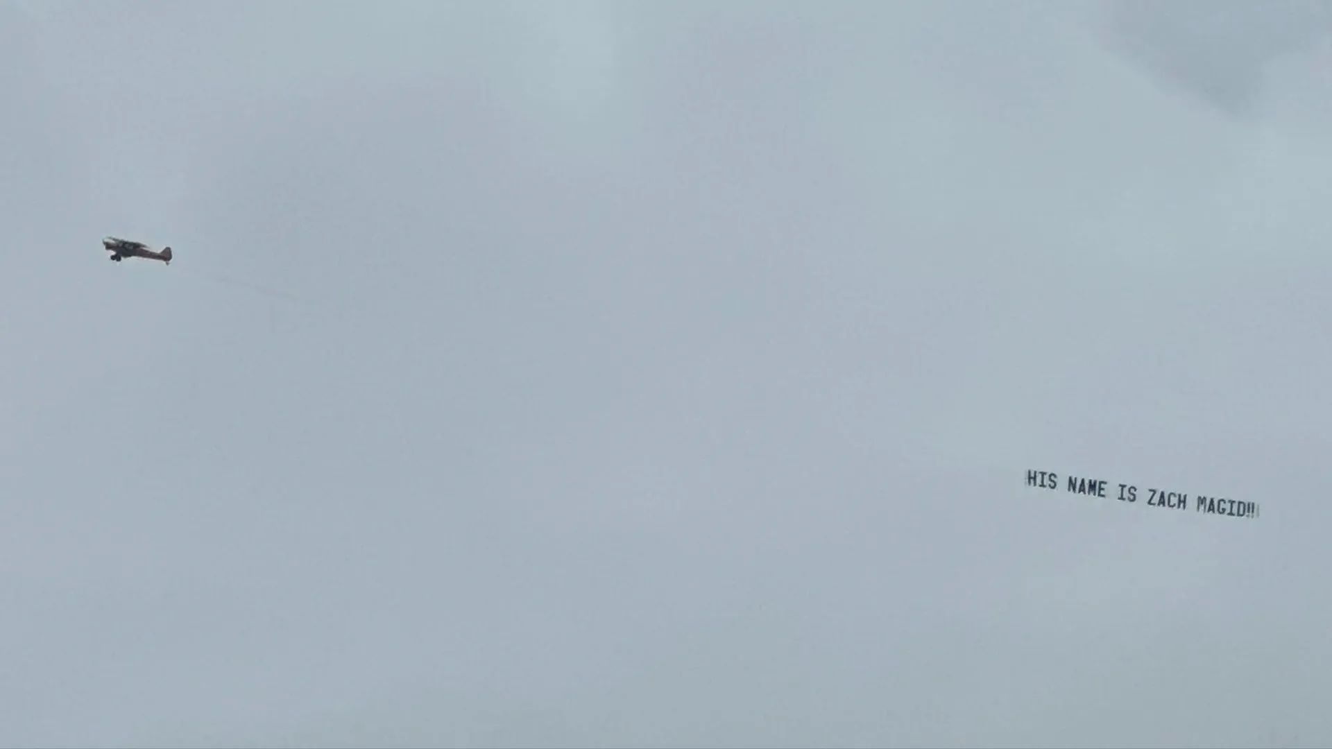 Small airplane flying in gray sky towing a banner that reads "HIS NAME IS ZACH MAGID!!" in black letters.
