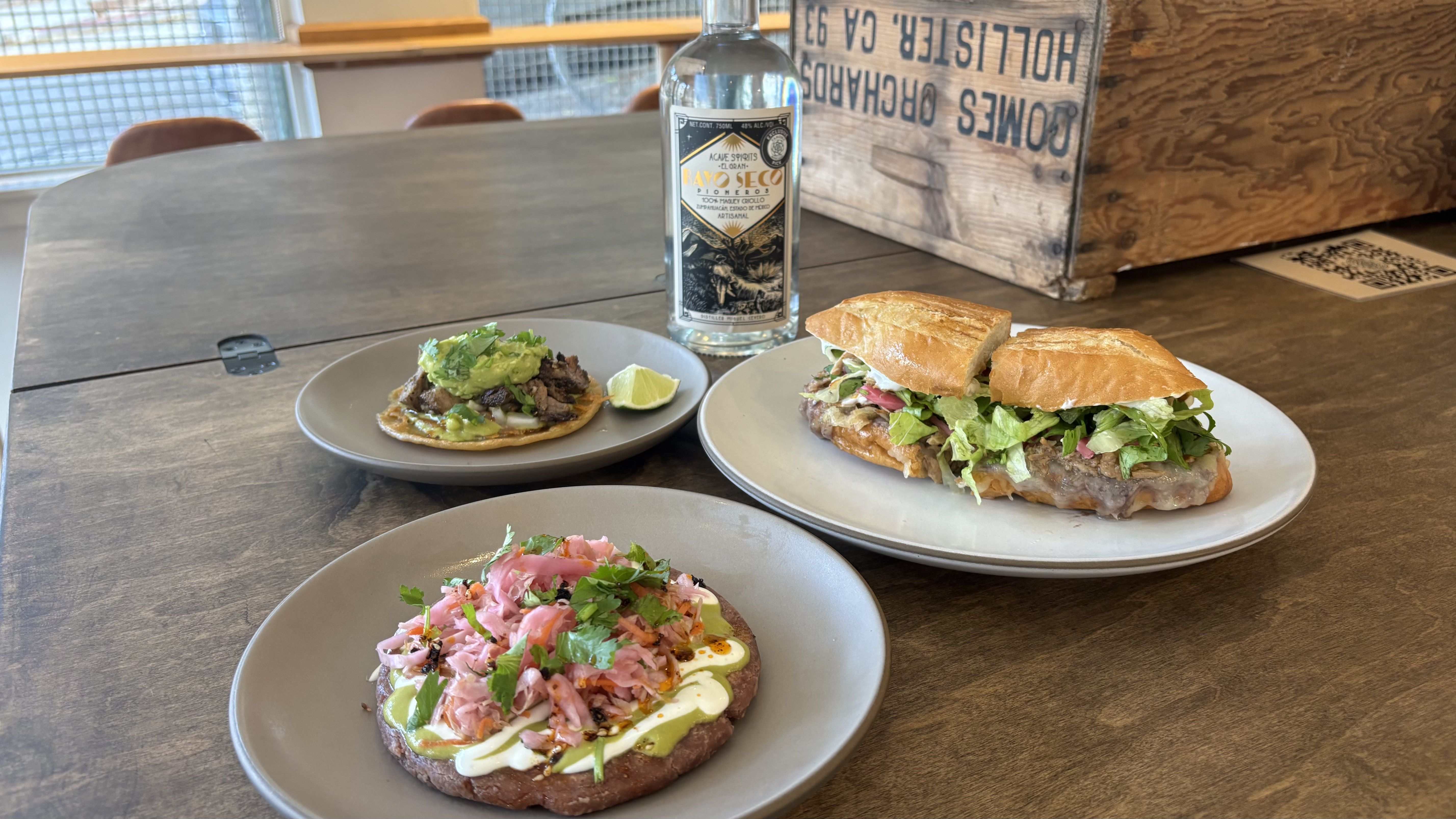 Three plates with Mexican dishes including a torta sandwich with lettuce, a taco with guacamole, and another dish with pink pickled vegetables, accompanied by a bottle of Rayo Seco agave spirit.