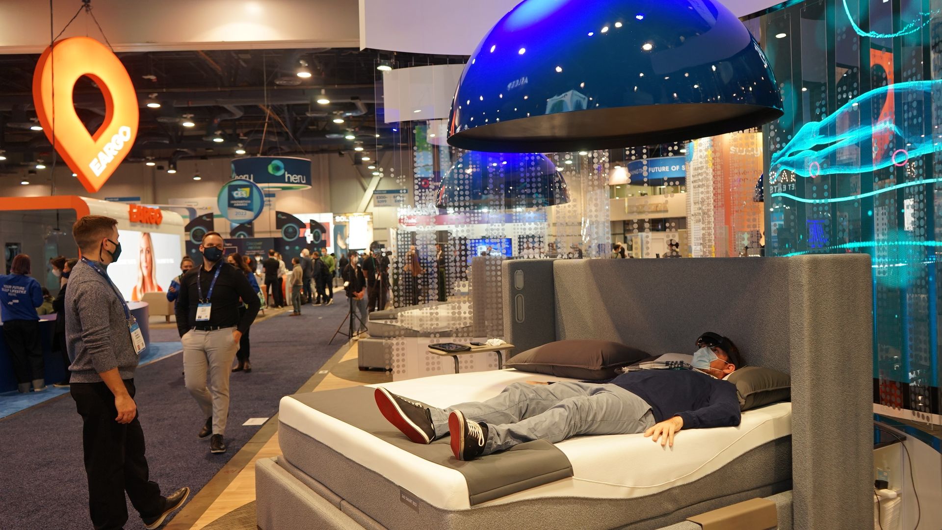 Person wearing a mask and VR headset lies on a gray bed with blue hanging lamps above at a busy tech expo with people and displays in the background.