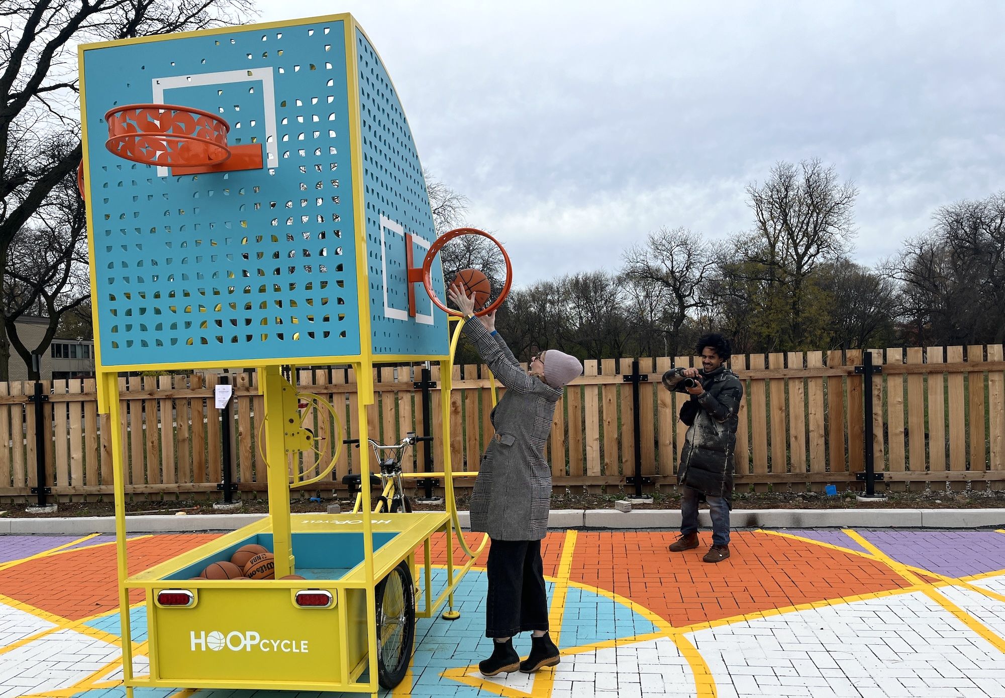 A woman in a hat and long coat puts a basketball through a vertical basketball rum on a blue basketball backboard on a yellow tricycle.