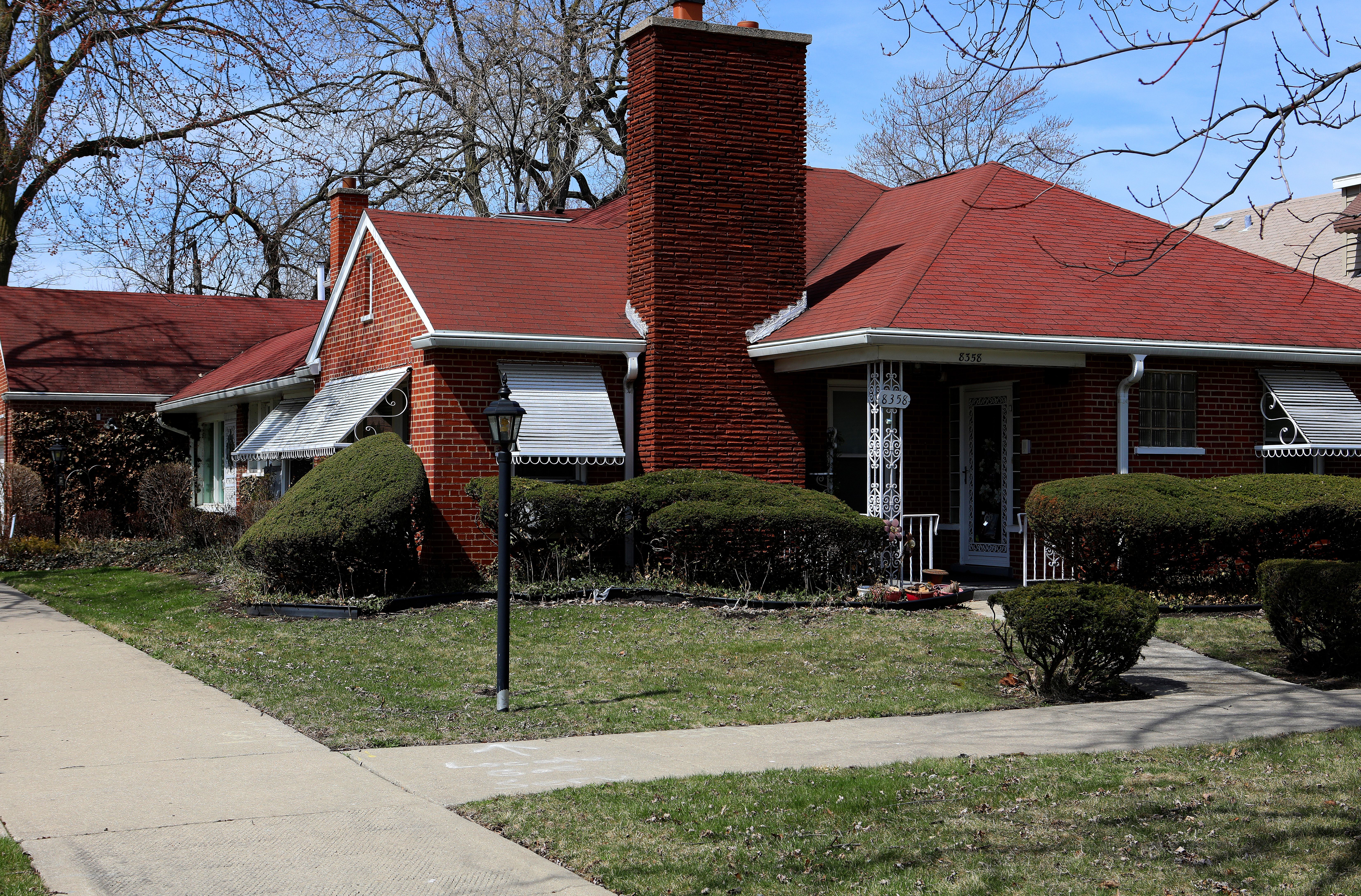 Single-story red brick house with a red shingle roof, large central chimney, white awnings, trimmed bushes, and black lamp posts in a grassy yard under a clear blue sky.