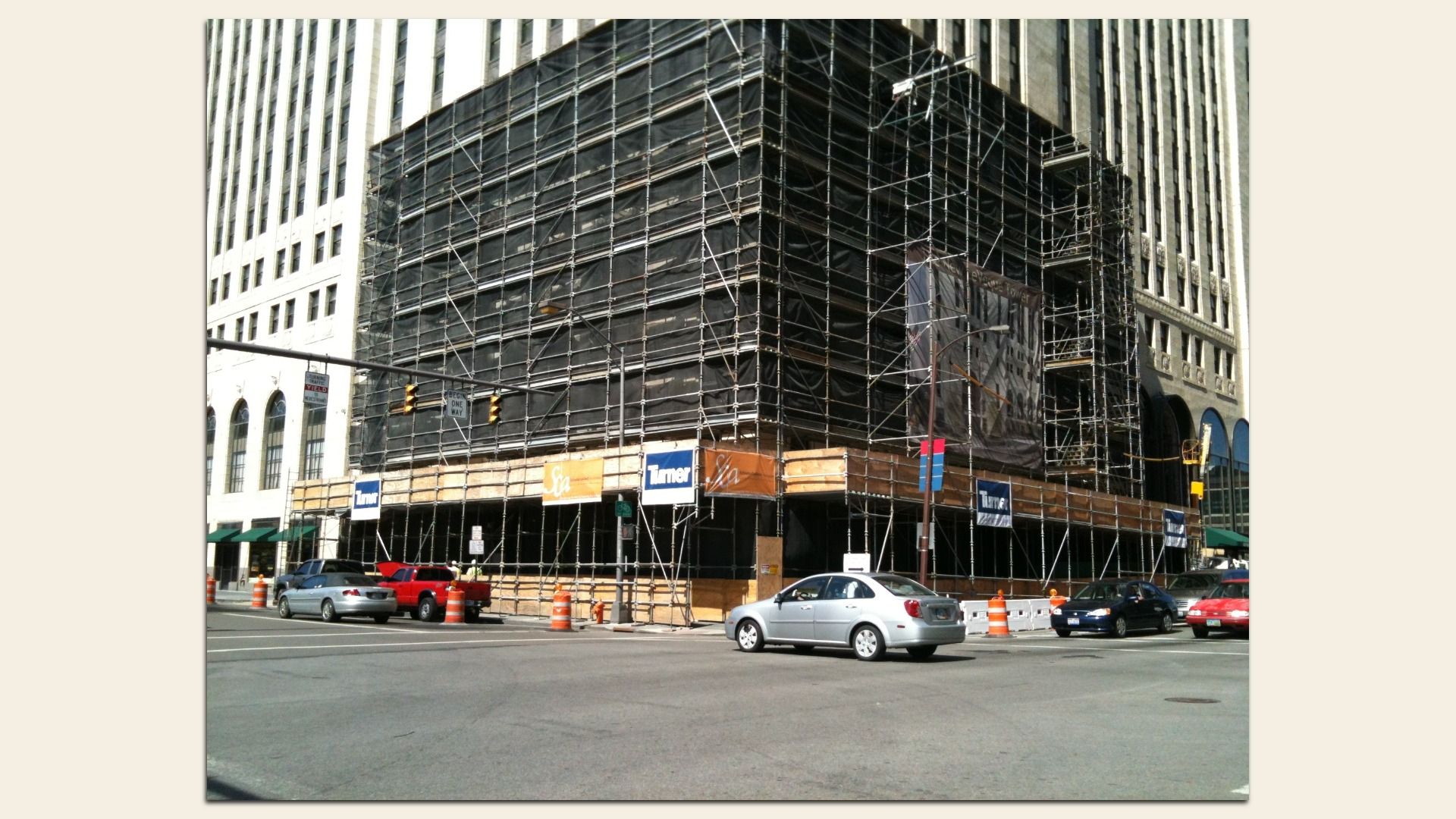 Construction site on a city street: a tall building wrapped in black netting and crisscrossed scaffolding, with orange traffic cones, parked cars, and banners at the base.