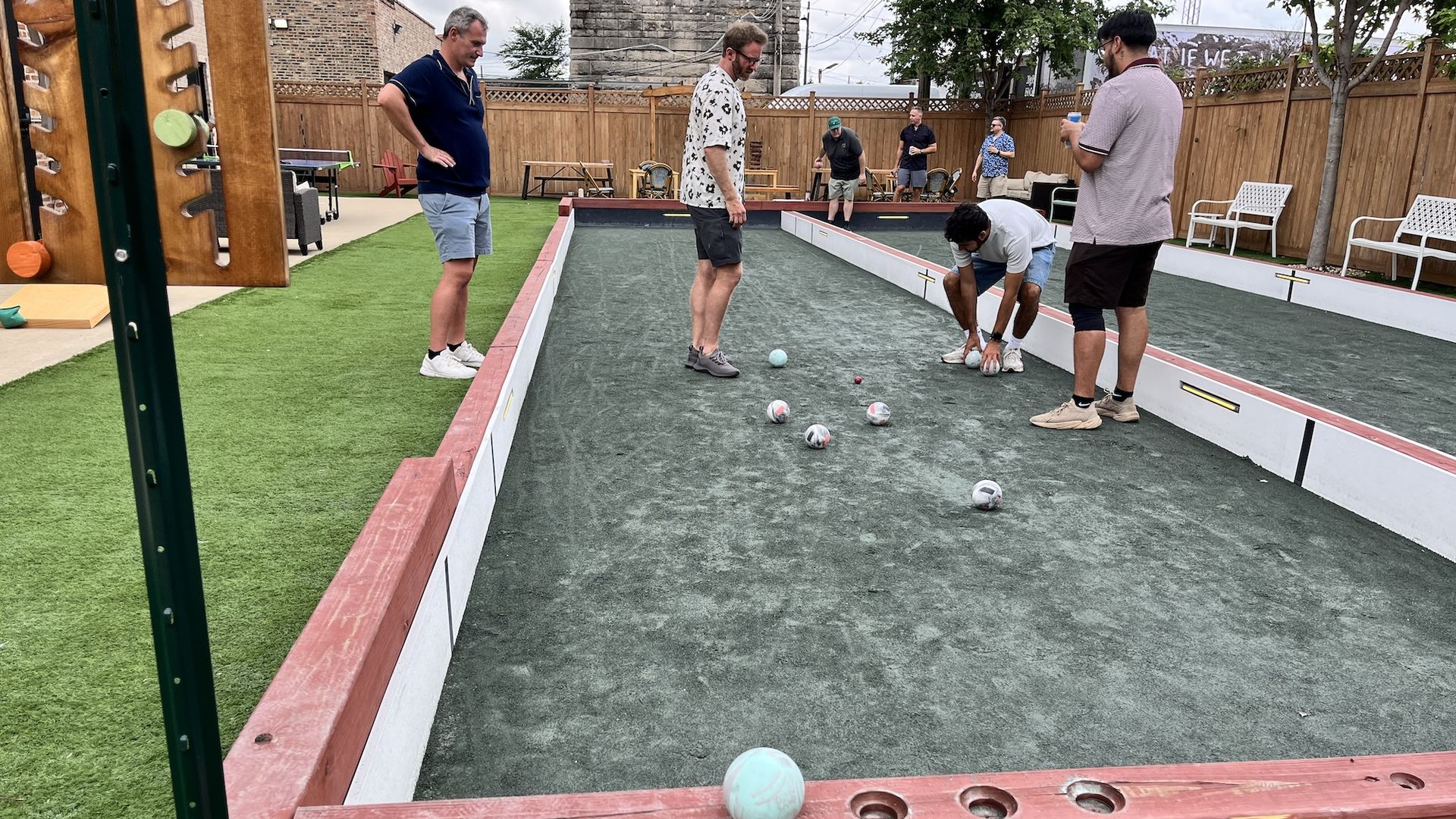 Several men stand around a small outdoor bocce court, with one bending to pick up a ball. Several balls scattered on the court; wooden fence, trees, and outdoor chairs in the background.