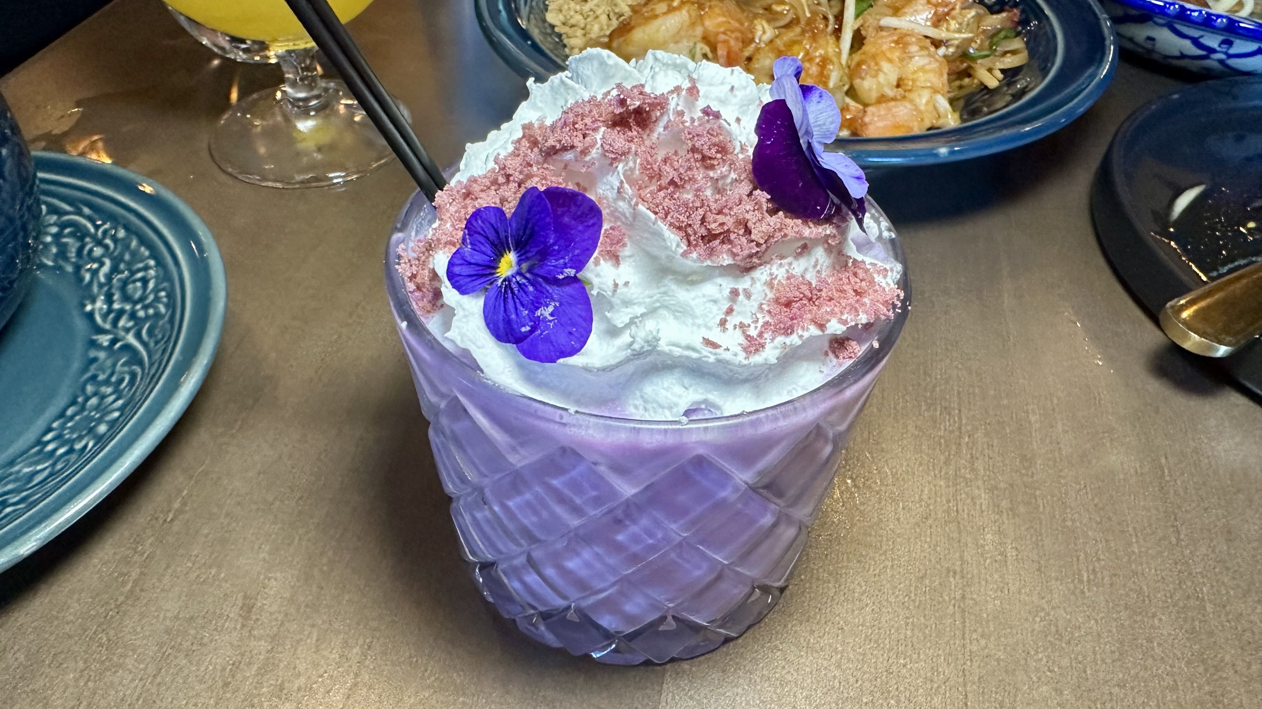 Purple ribbed glass holds a dessert topped with white whipped cream, pink crumble, and two purple edible flowers. A black straw protrudes; teal plate and a bowl of food are in the background.