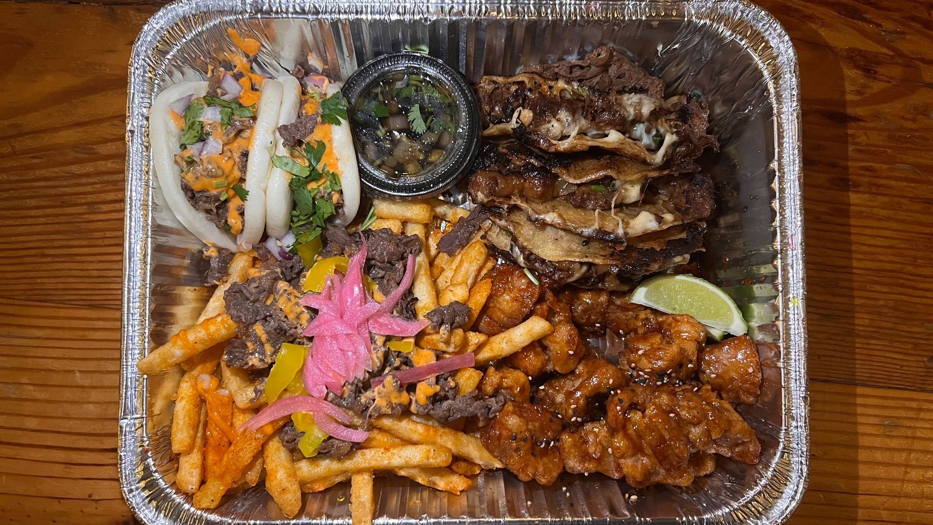 A picture of tray with bao buns on the top left corner, then sauce, then birria tacos, some limes, chicken and seasoned fries topped with beef and pickled onions on the bottom left