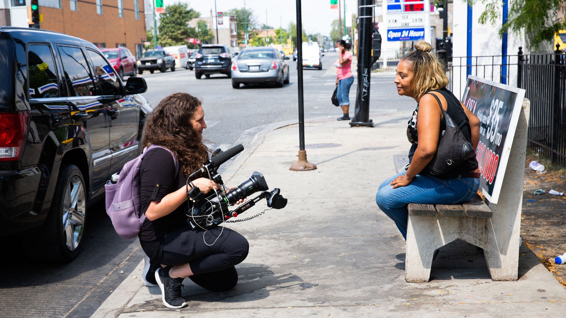 A woman interviews another woman at a bus stop.