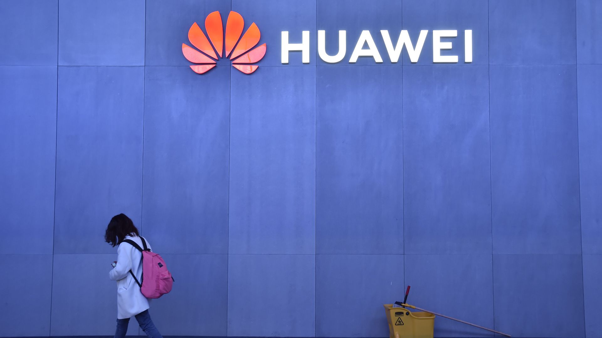 A blue wall with a neon sign that says "Huawei." There is a young person wearing a backpack walking in front of the wall.
