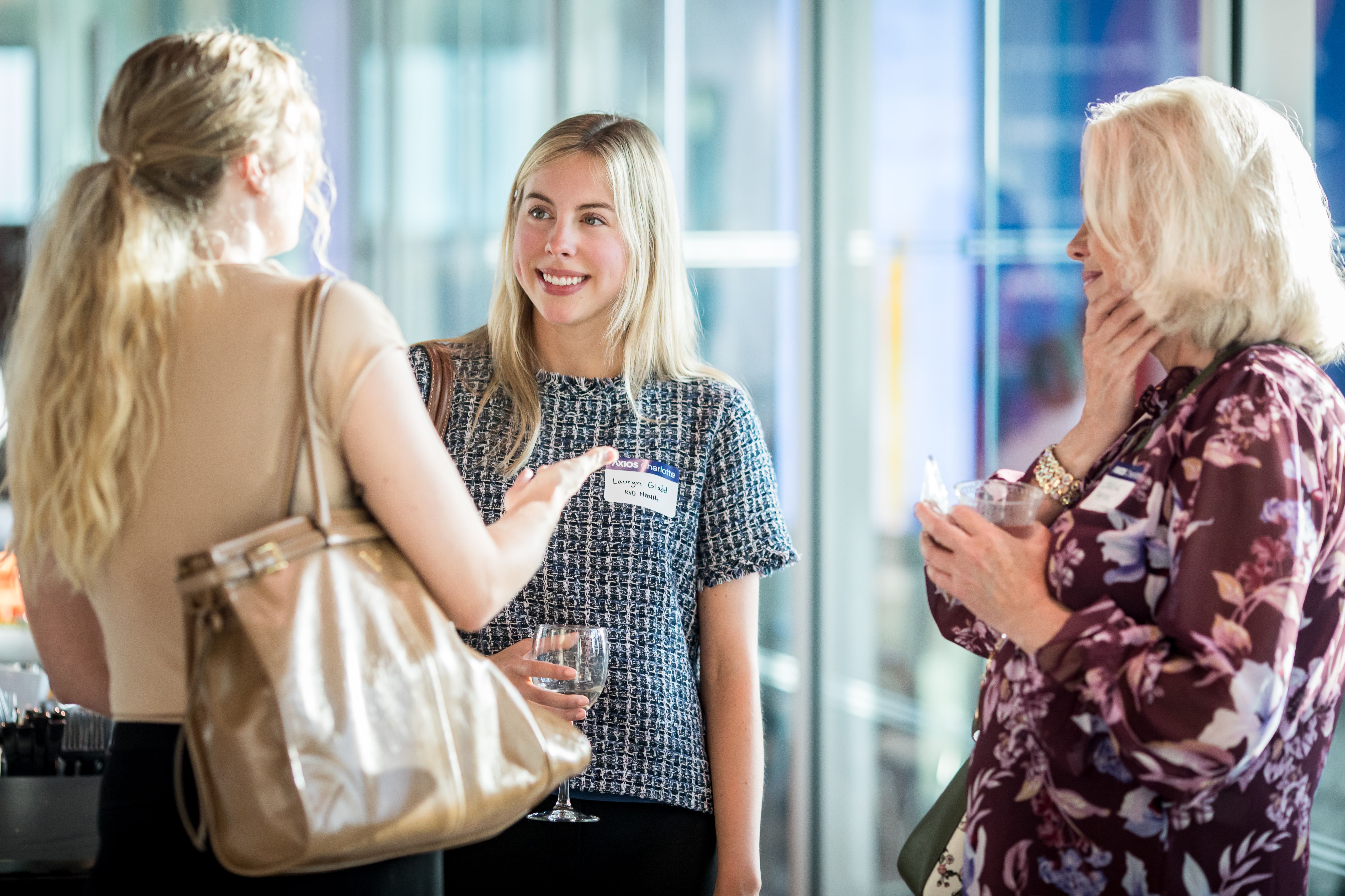Three women chat at a social event by glass walls; center blonde with name badge smiles, holding a wine glass, flanked by a blonde on the left and an older woman in a purple floral dress on the right.