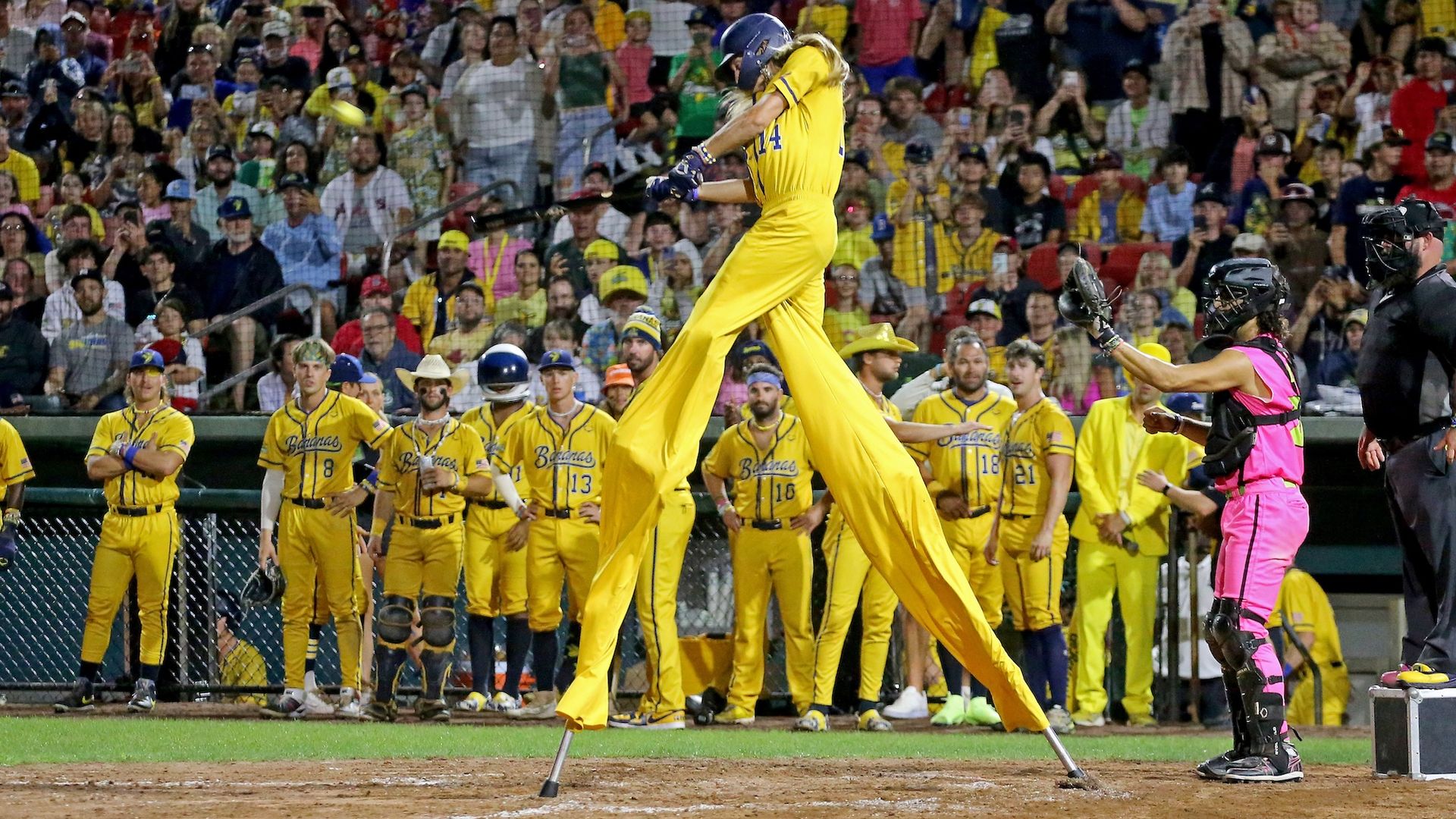 A baseball batter in a bright yellow uniform swings mid-air as the ball approaches; a colorful crowd roars in the stands while yellow-clad teammates and a pink-vested umpire watch.