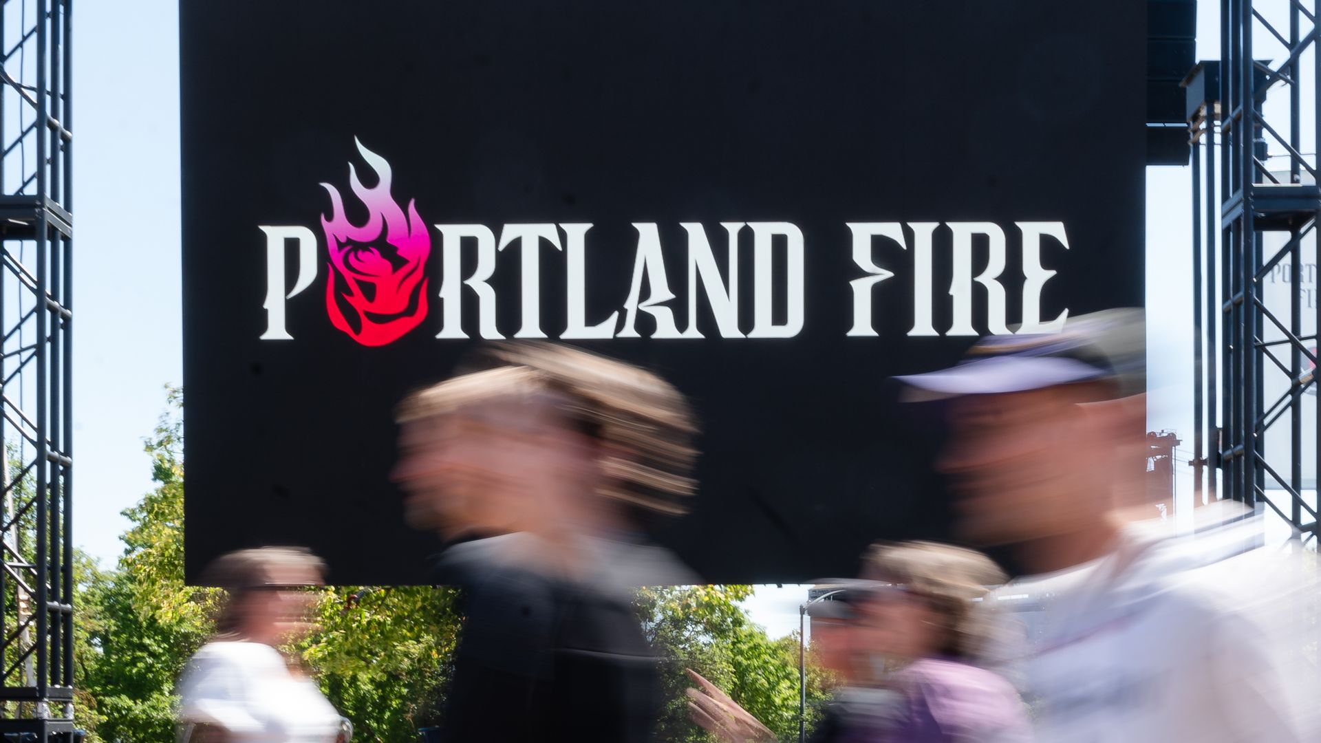 Blurred pedestrians walk in front of a large black billboard reading "PORTLAND FIRE" with a pink and red flame logo to the left; outdoor scene with green trees and metal scaffolding on the sides.