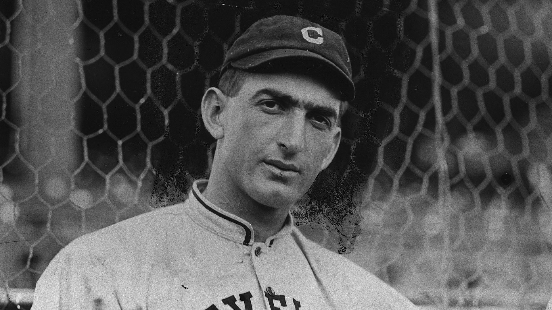 "Shoeless" Joe Jackson stands in front of a fence in a Cleveland baseball uniform. 