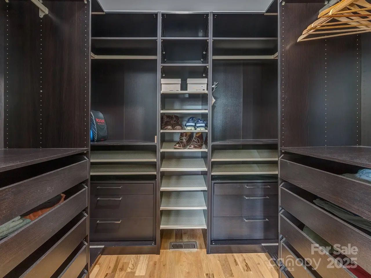 Dark wood walk-in closet with multiple drawers, shelves holding shoes and boxes, and some folded clothes, resting on a medium-tone hardwood floor.
