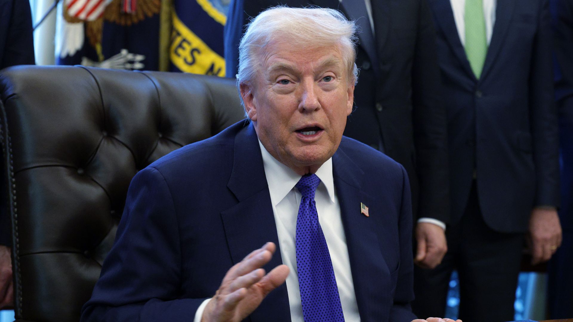 President Trump with white hair in a navy suit and blue tie sitting at a desk, gesturing with his right hand. American flag and officials in suits stand behind him.