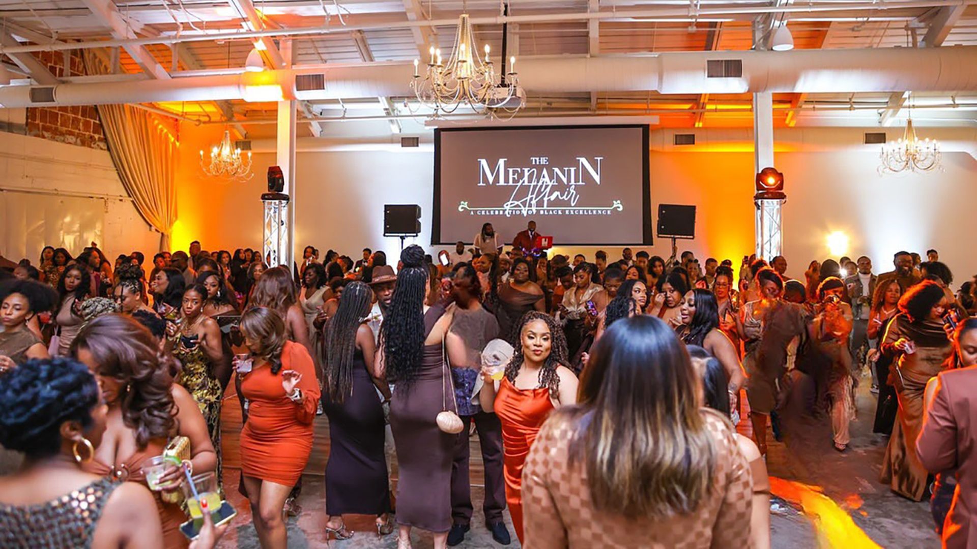 Crowd of people dressed in elegant warm tones at an indoor event with chandeliers and a screen reading "The Melanin Affair: A Celebration of Black Excellence" in orange and white lighting.