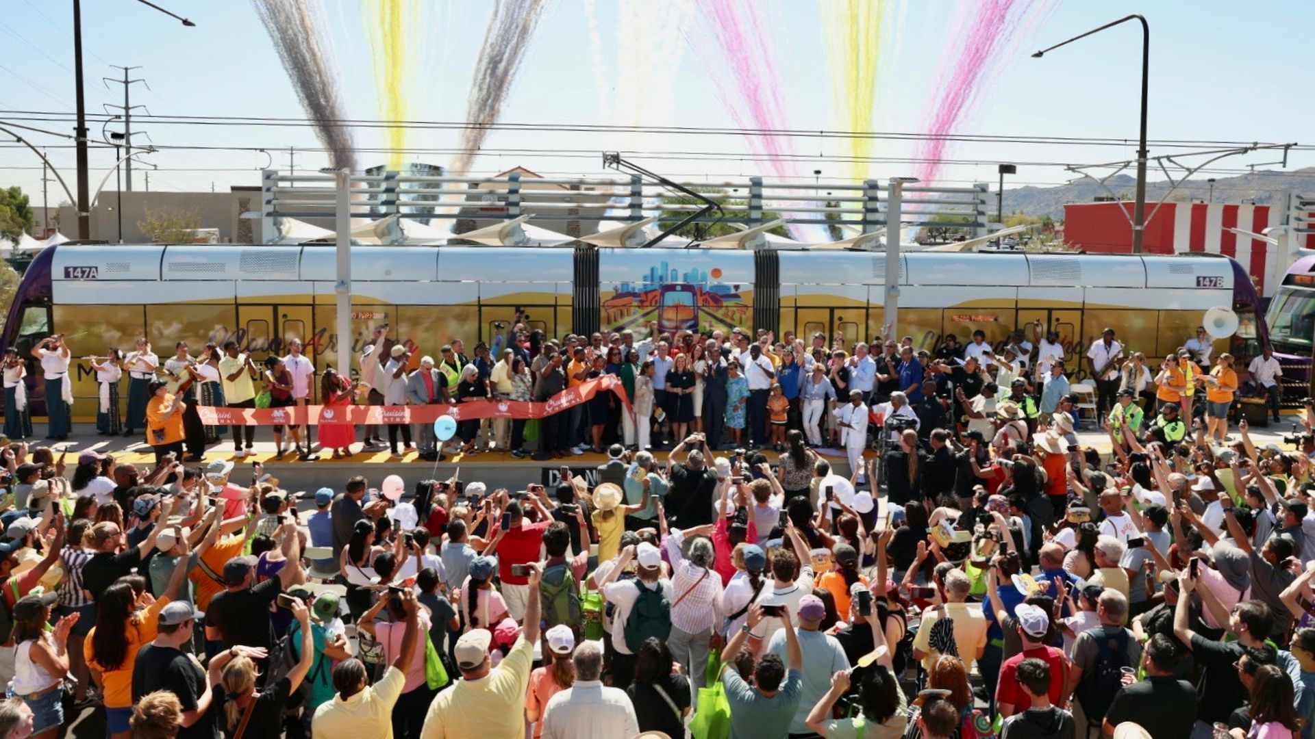 A large crowd is gathered in front of a light rail train platform where officials had just cut a large red ribbon. 