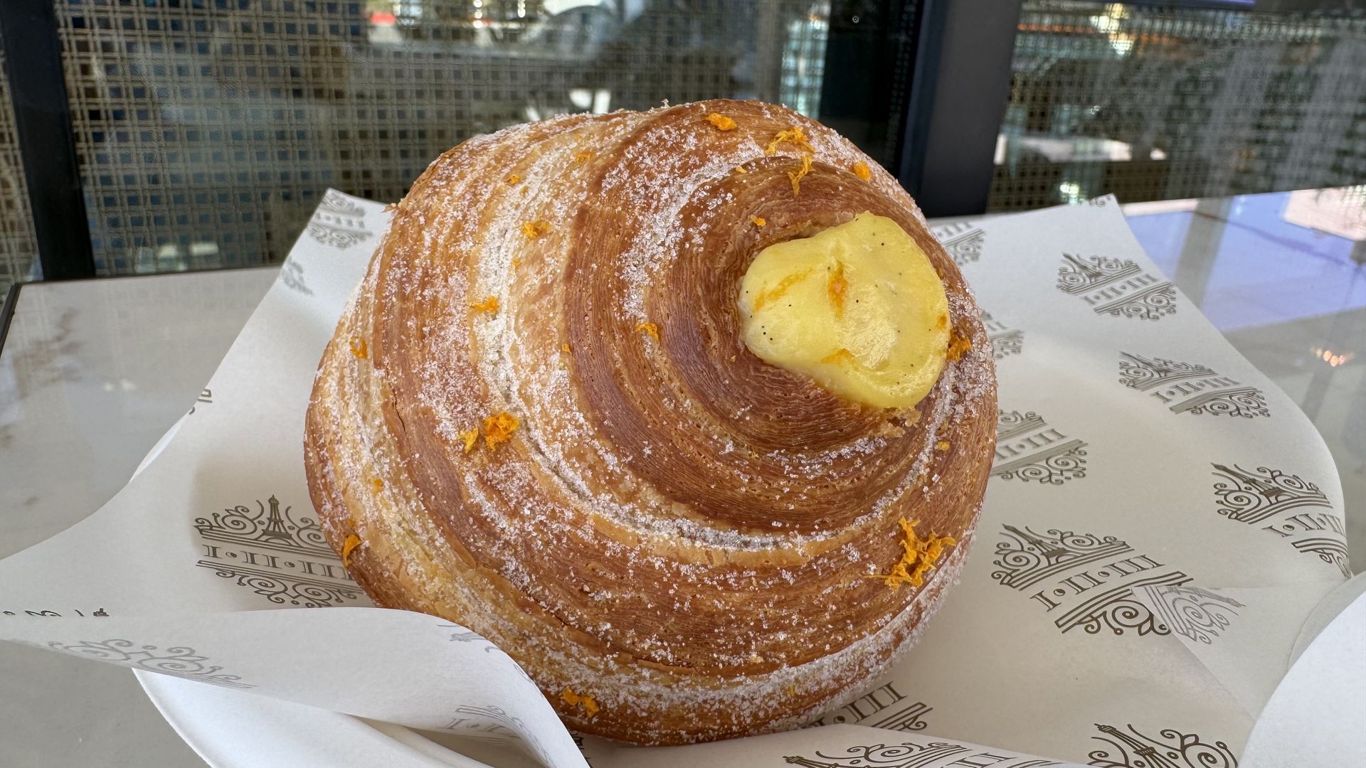 A golden, sugar-dusted spiral pastry filled with yellow custard sits on decorative bakery paper at a marble-topped table.