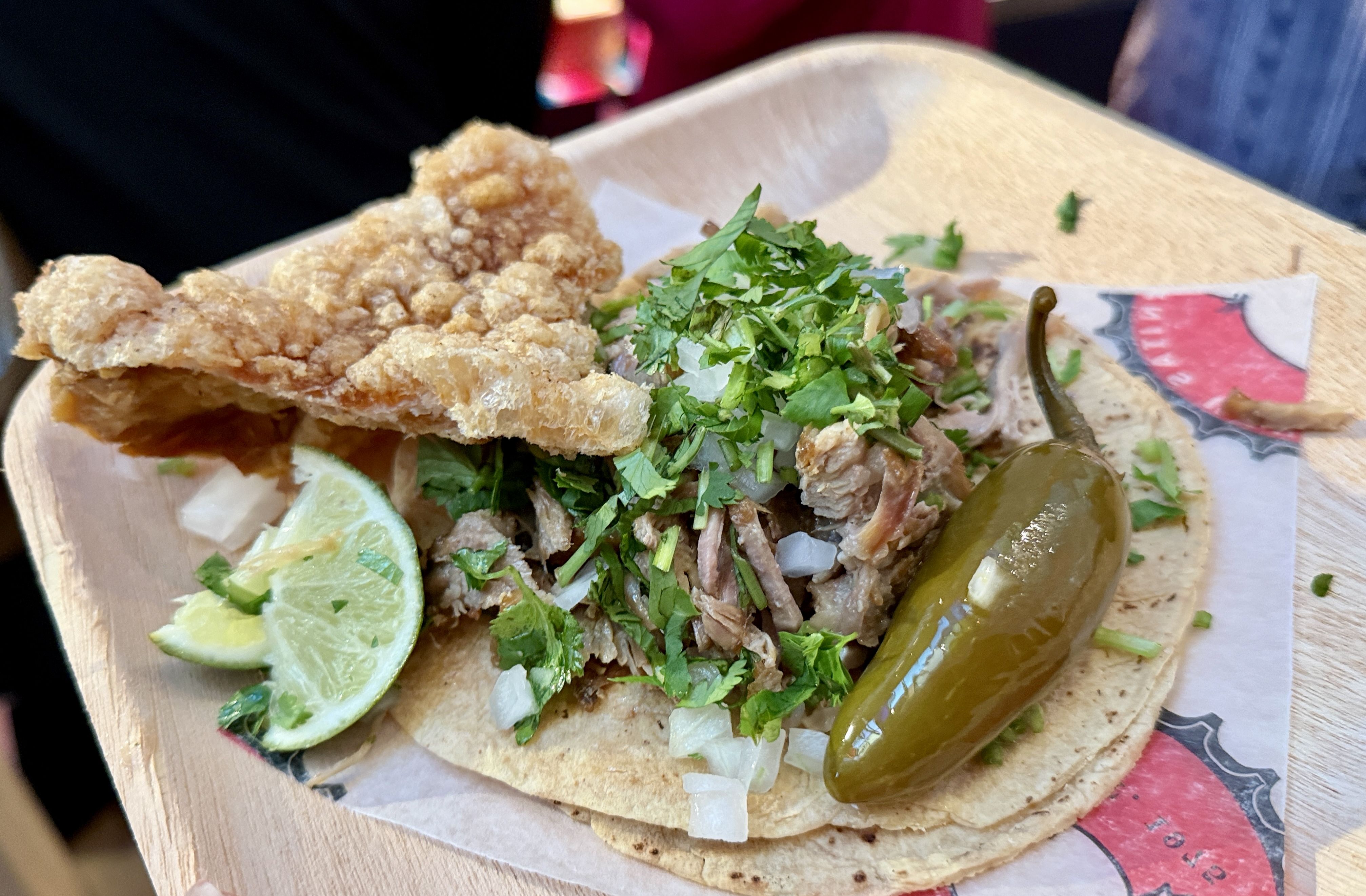 Close-up of a street-style taco on a wooden plate: crispy fried chicken, chopped onions, fresh cilantro, a green jalapeño, and lime wedges.