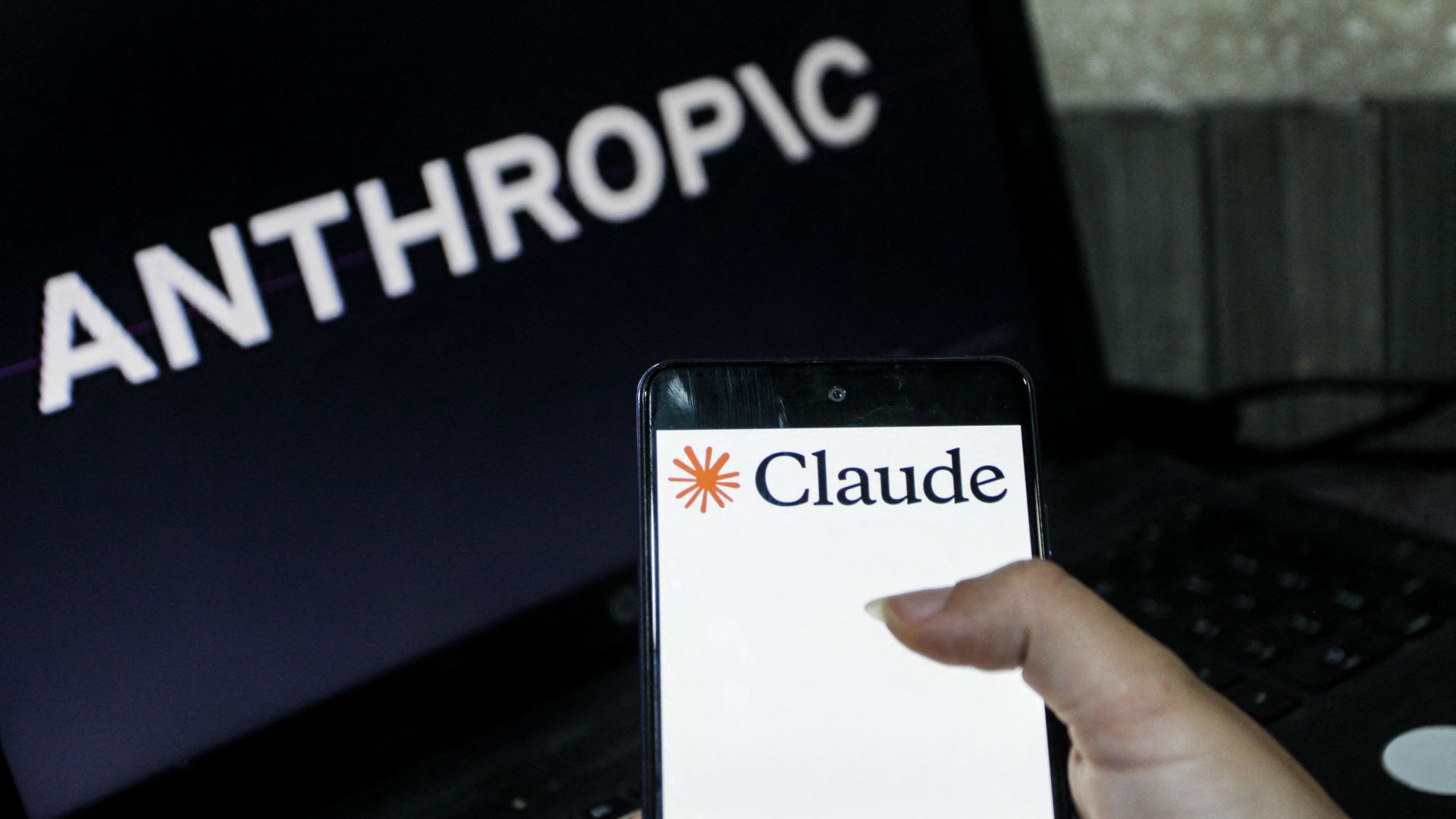Close-up of a laptop screen with 'ANTHROPIC' in white on a dark background; in front, a hand holds a smartphone showing a badge that says 'Claude' with an orange sunburst logo.