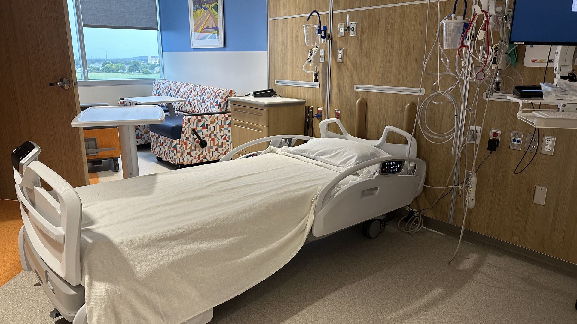 Hospital room with two beds, white linens, and tangled medical cords along a wood-paneled wall. A colorful patterned chair sits by a window with a blue wall and a painting.