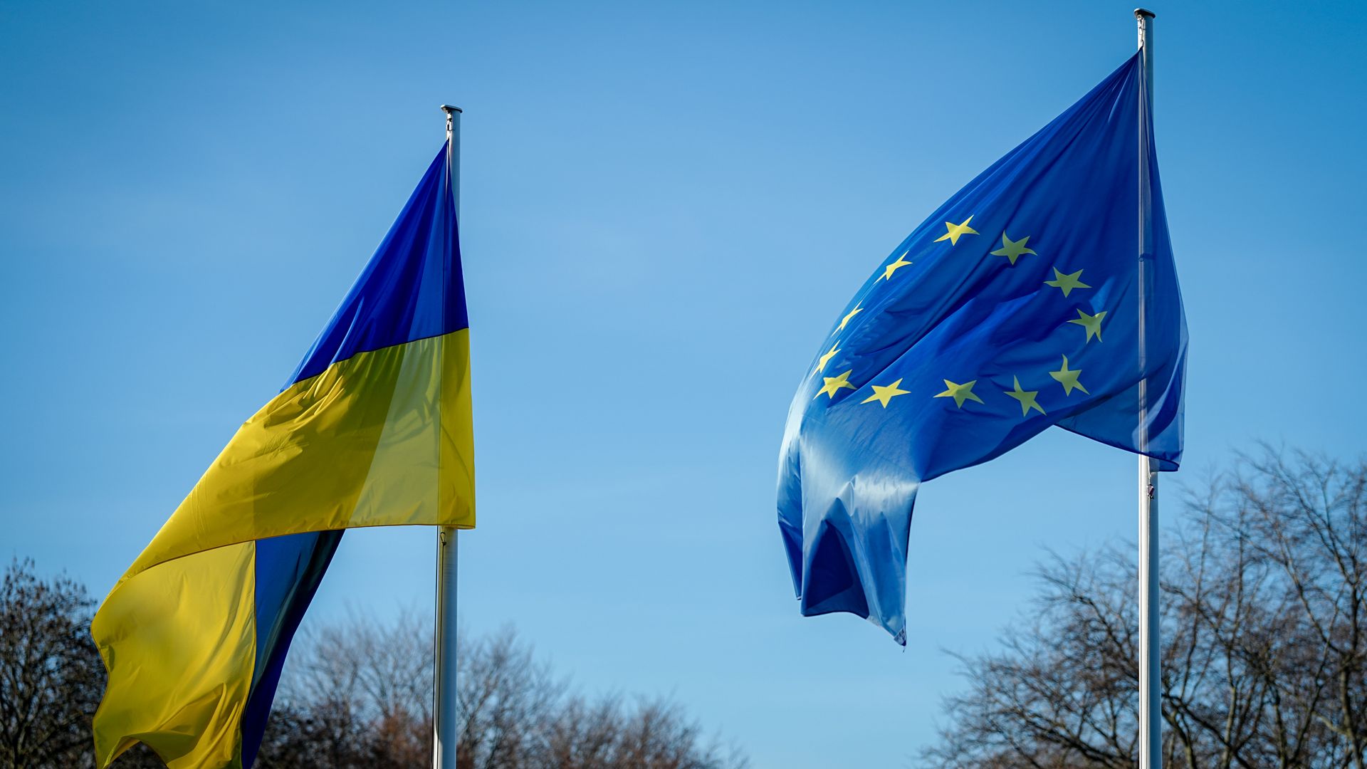 Two flags waving on flagpoles against a clear blue sky and bare trees: the Ukraine flag on the left with blue and yellow horizontal stripes, and the European Union flag on the right with yellow stars in a circle on blue.
