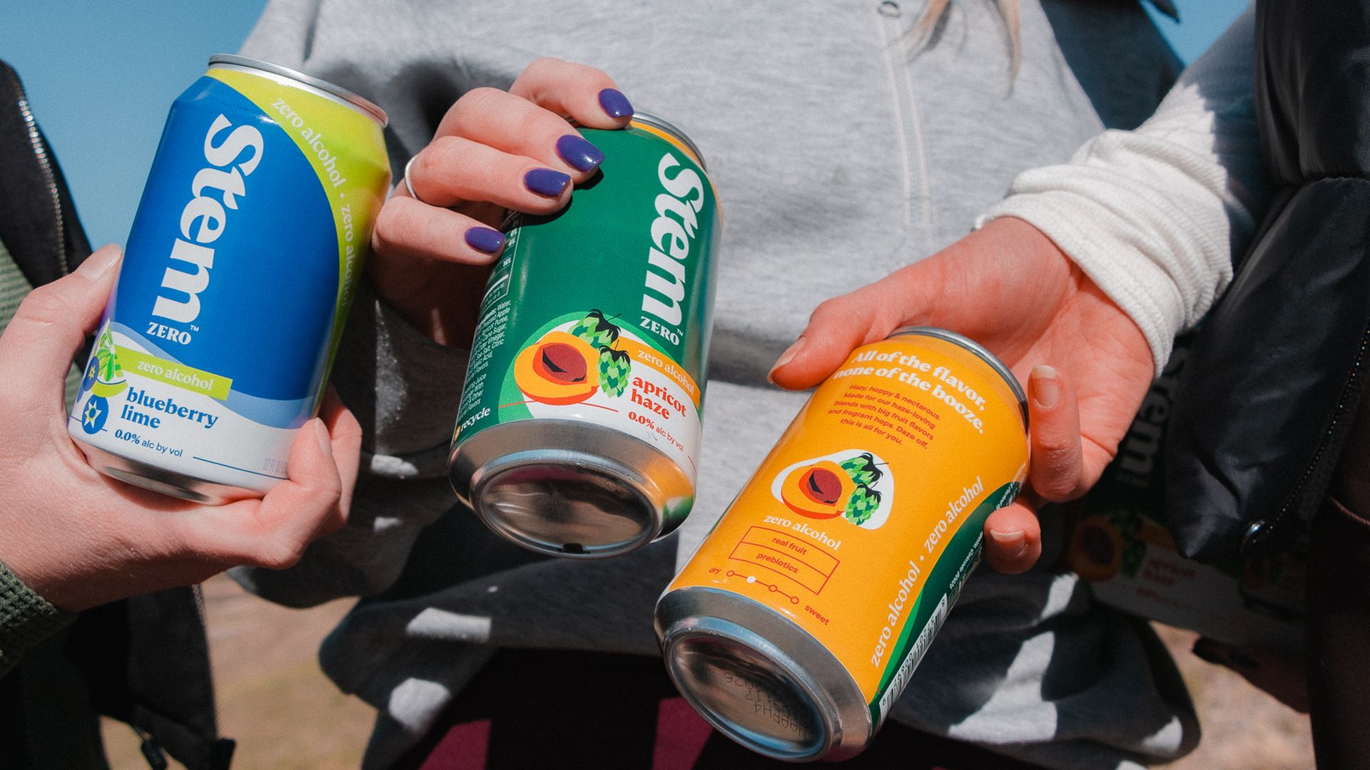 Three people holding colorful cans of Stem zero alcohol beverages outdoors, flavors visible include blueberry lime and apricot haze.