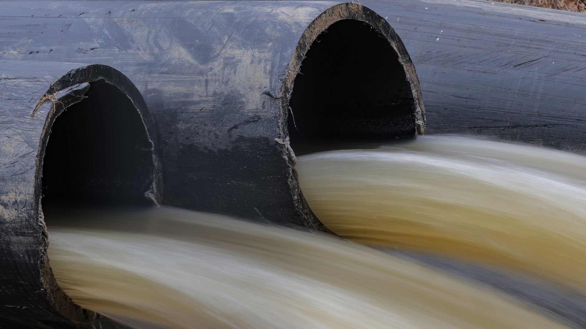 Water flowing rapidly out of two large black drainage pipes, with blurred motion capturing the flow and some fallen leaves visible on the ground near the pipes.