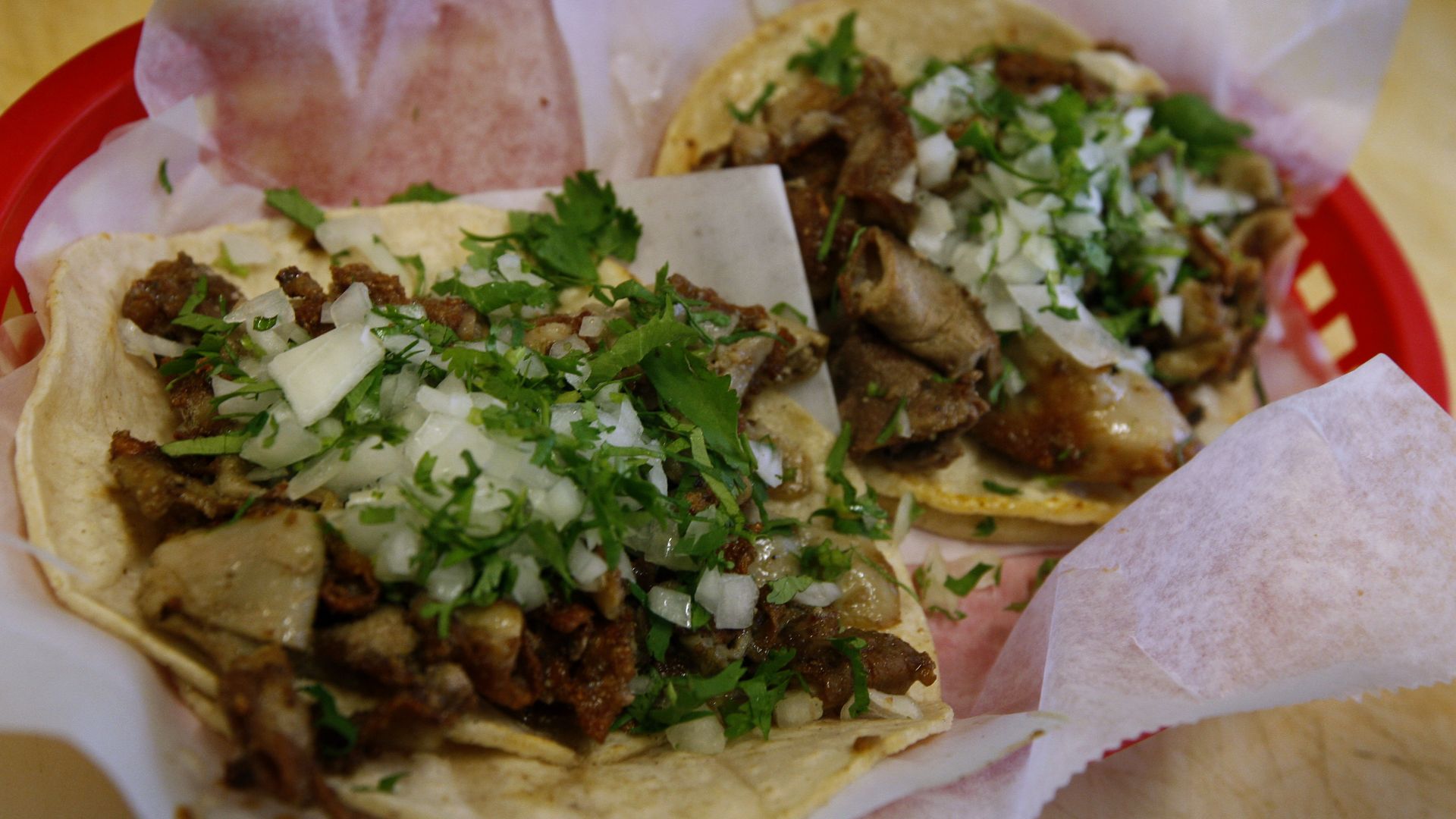 Two tacos in a red basket lined with white paper, filled with seasoned meat, chopped white onions, and fresh green cilantro.