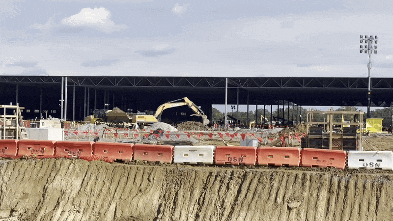 An excavator digs into a pile of dirt, viewed from afar. Behind it, a large under-construction steel-frame building.