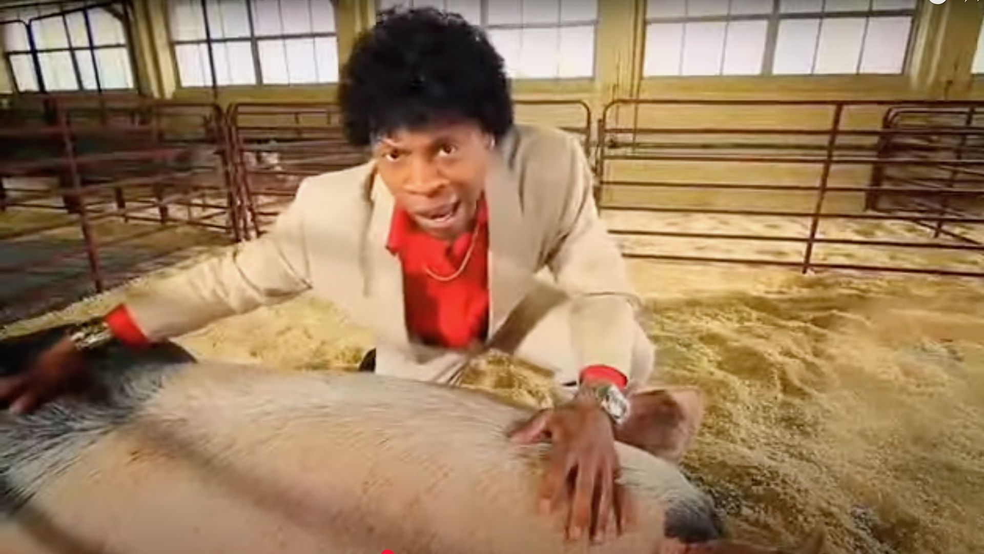 An actor in a beige suit pets a pig in a barn stall. 