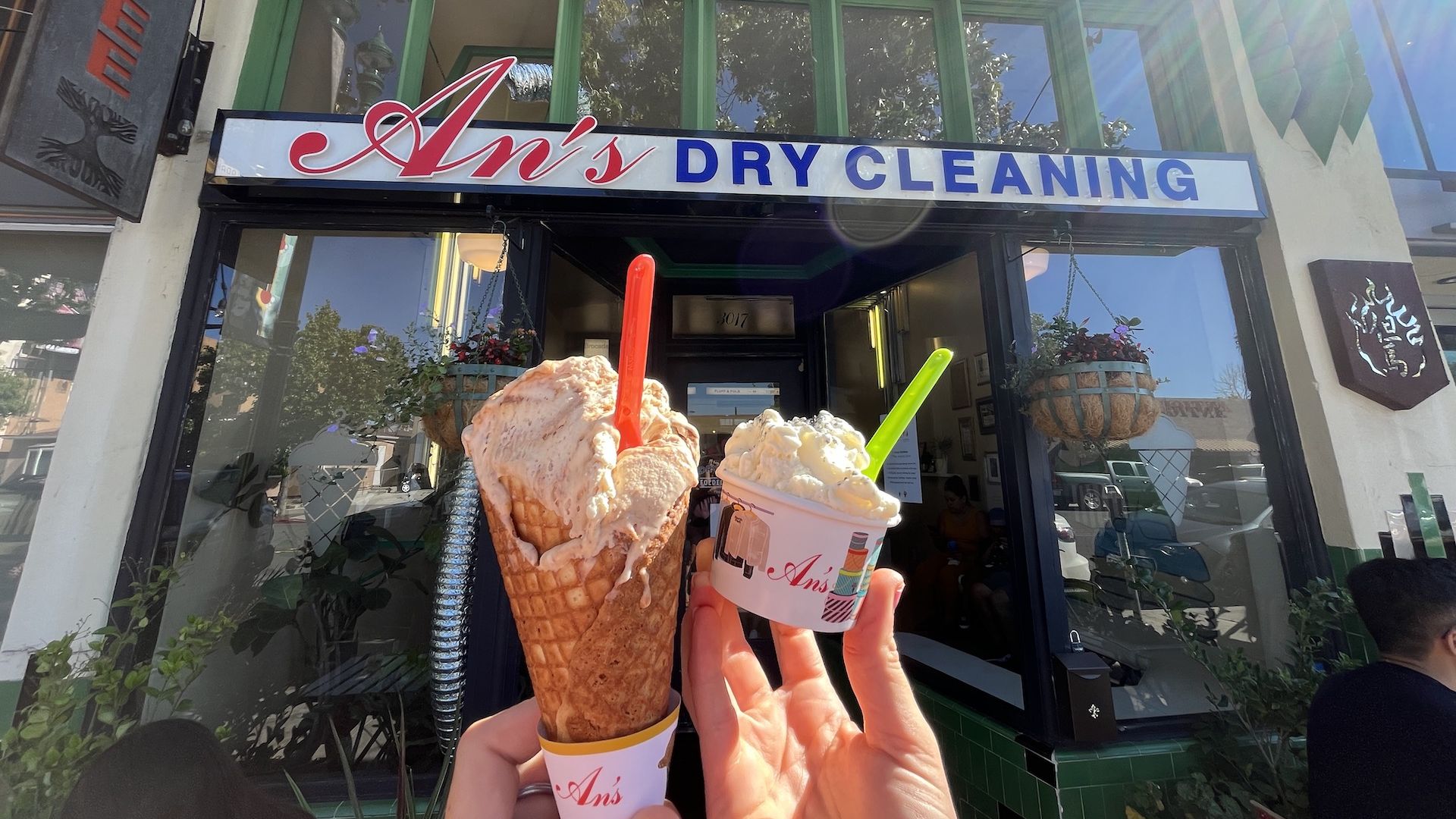Two hands hold up scoops of ice cream in a cone and a cup in front of a shop that says An's dry cleaning.