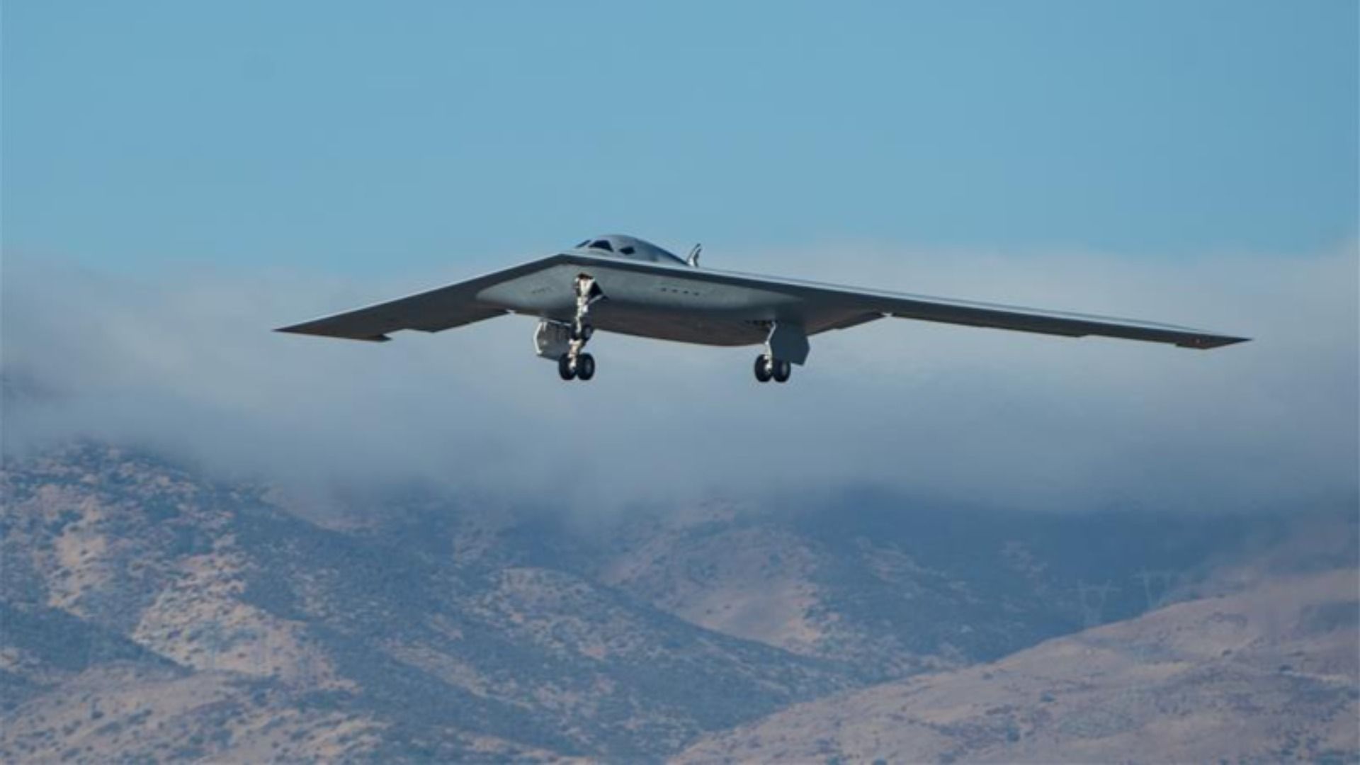 A gray B-21 Raider stealth bomber flies low over a rugged mountain landscape.